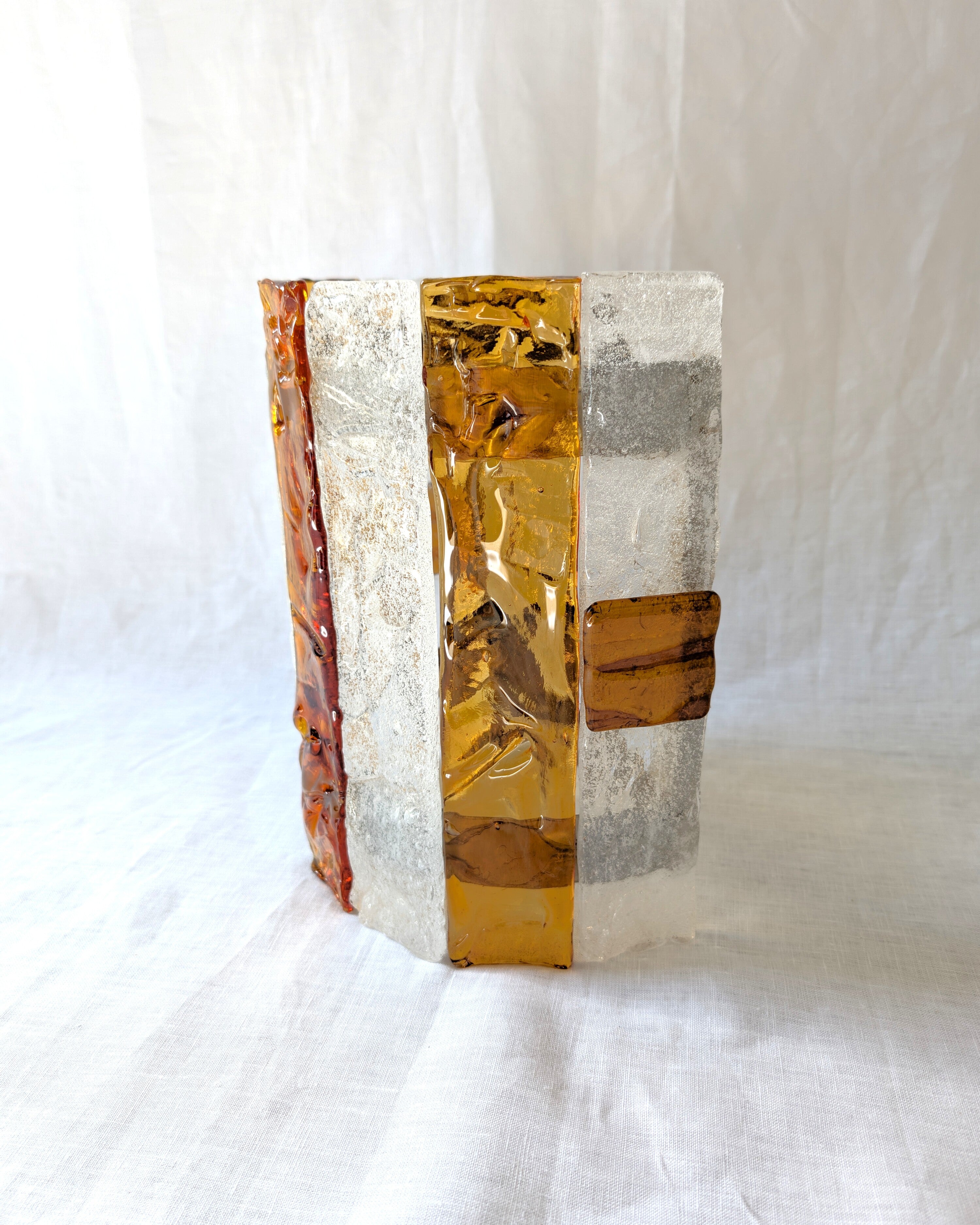 The side of a vintage mid century modern 1970s Carlo Nason for Mazzega rectangular wall sconce with textured amber and clear striped Murano glass panels on white linen surface.