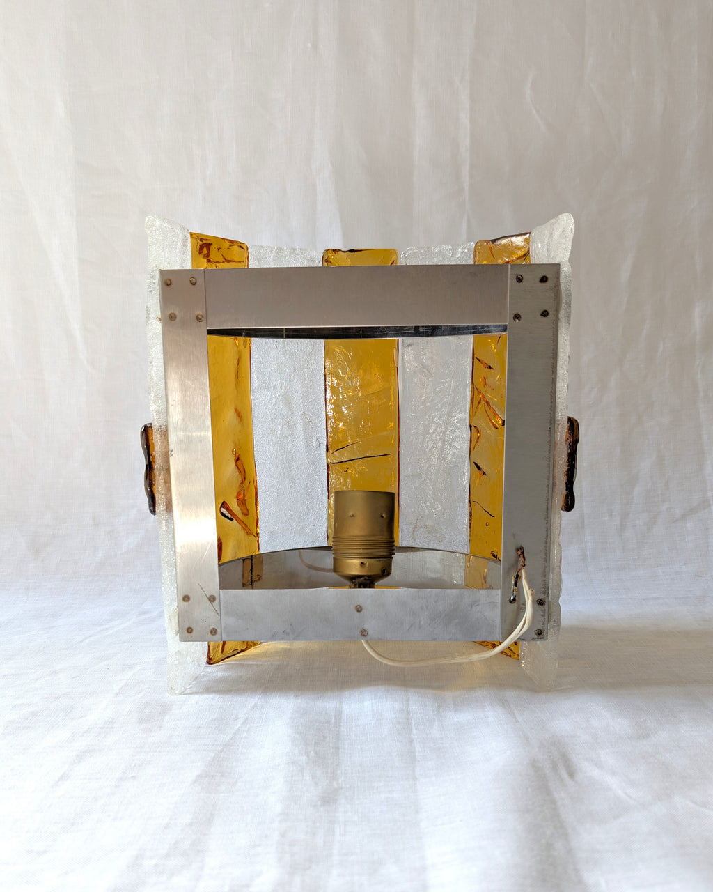 The back of a vintage mid century modern 1970s Carlo Nason for Mazzega rectangular wall sconce showing the metal hanging bracket, light bulb socket and wiring, with textured amber and clear striped Murano glass panels on white linen surface.