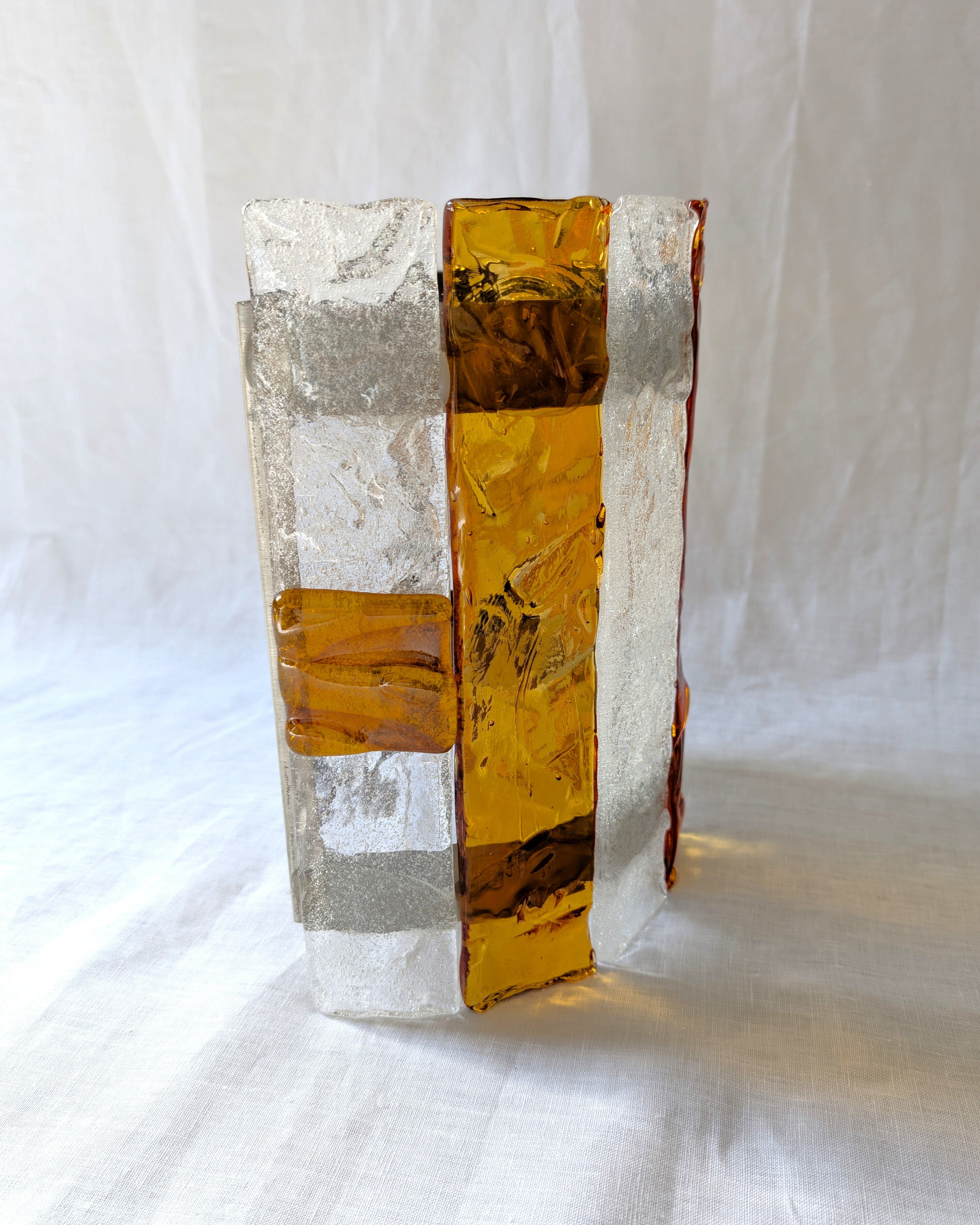 A close up of the side of a vintage mid century modern 1970s Carlo Nason for Mazzega rectangular wall sconce showing the metal hanging bracket, light bulb socket and wiring, with textured amber and clear striped Murano glass panels on white linen surface.