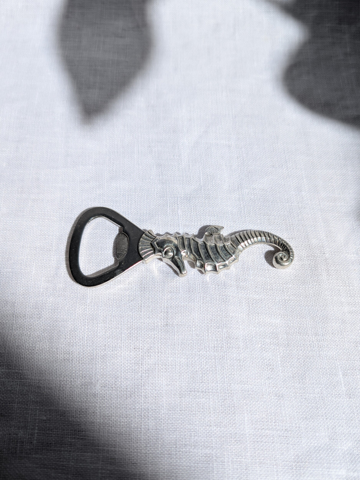 A vintage 1930s art deco Just Andersen pewter seahorse shaped bottle opener sitting horizontally on white linen with shadows cast on it. This Danish mid century modern bottle opener features a loop for leverage, a detailed seahorse carved design, a curved tail and the Just Andersen makers engraving of 'Just Denmark' on the fin to certify authenticity. A wonderful quirky addition to a retro bar cart.