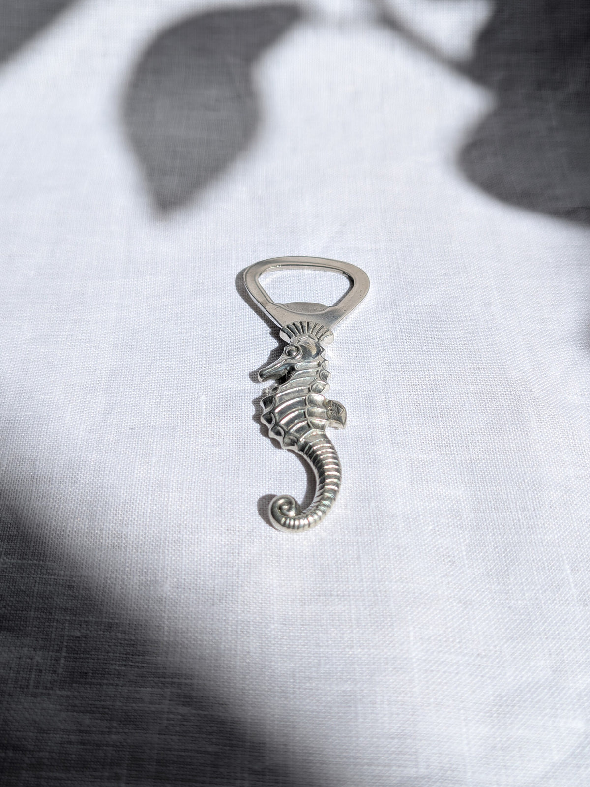 A vintage 1930s art deco Just Andersen pewter seahorse shaped bottle opener on white linen with shadows cast on it. This Danish mid century modern bottle opener features a loop for leverage, a detailed seahorse carved design, a curved tail and the Just Andersen makers engraving of 'Just Denmark' on the fin to certify authenticity. A wonderful quirky addition to a retro bar cart.