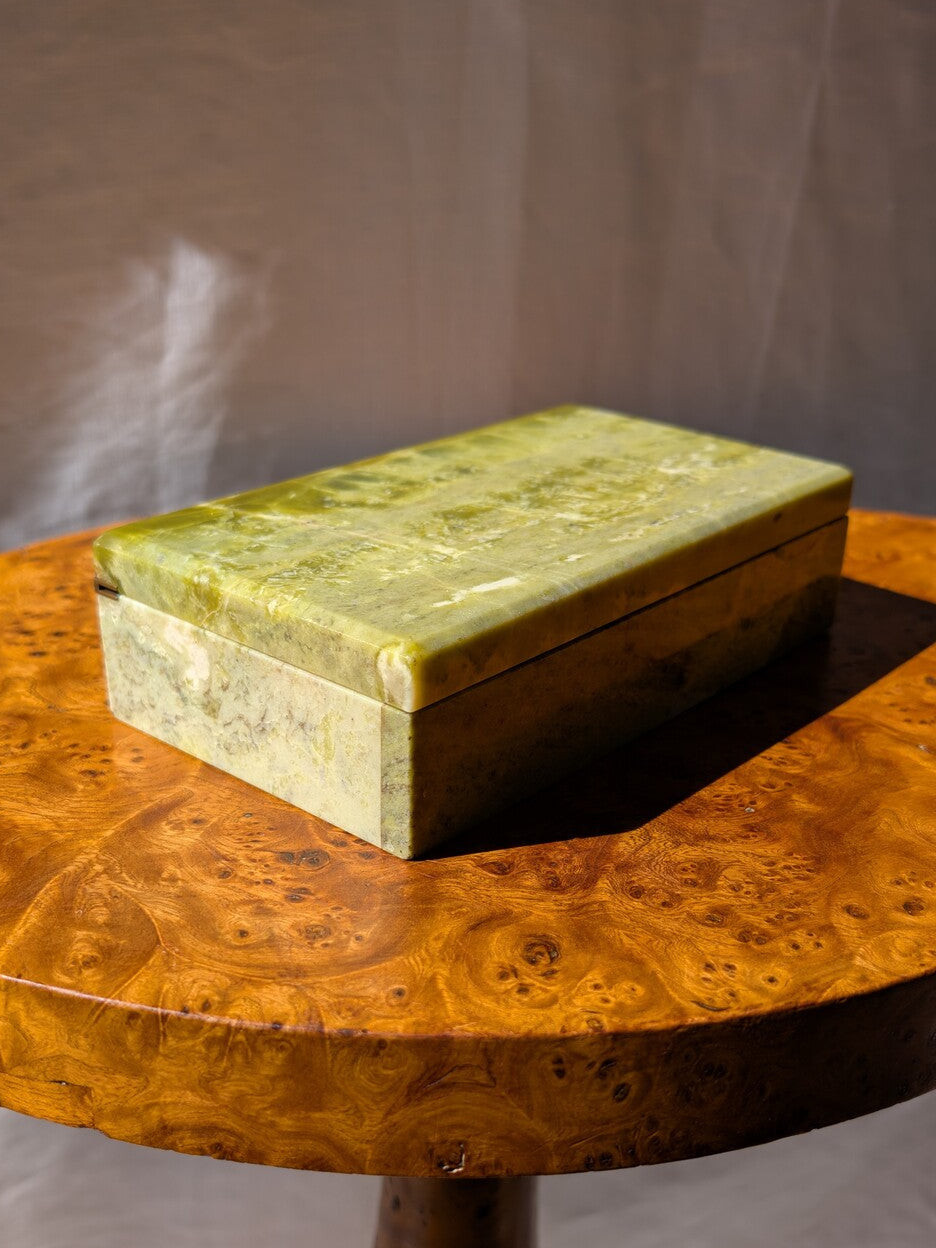 The side view of an antique green marble decor box by Henry Griffith & Sons, open to show the inside, on a Hollywood Regency style burl wood table. This rare Victorian decor box, made from hand carved solid Italian marble similar to onyx or jade, can be used as a trinket box, jewellery holder or cigar box and would be a beautiful addition to a vintage eclectic, mid century modern, minimalist, modernist, maximalist, baroque or art deco home interior schemes.