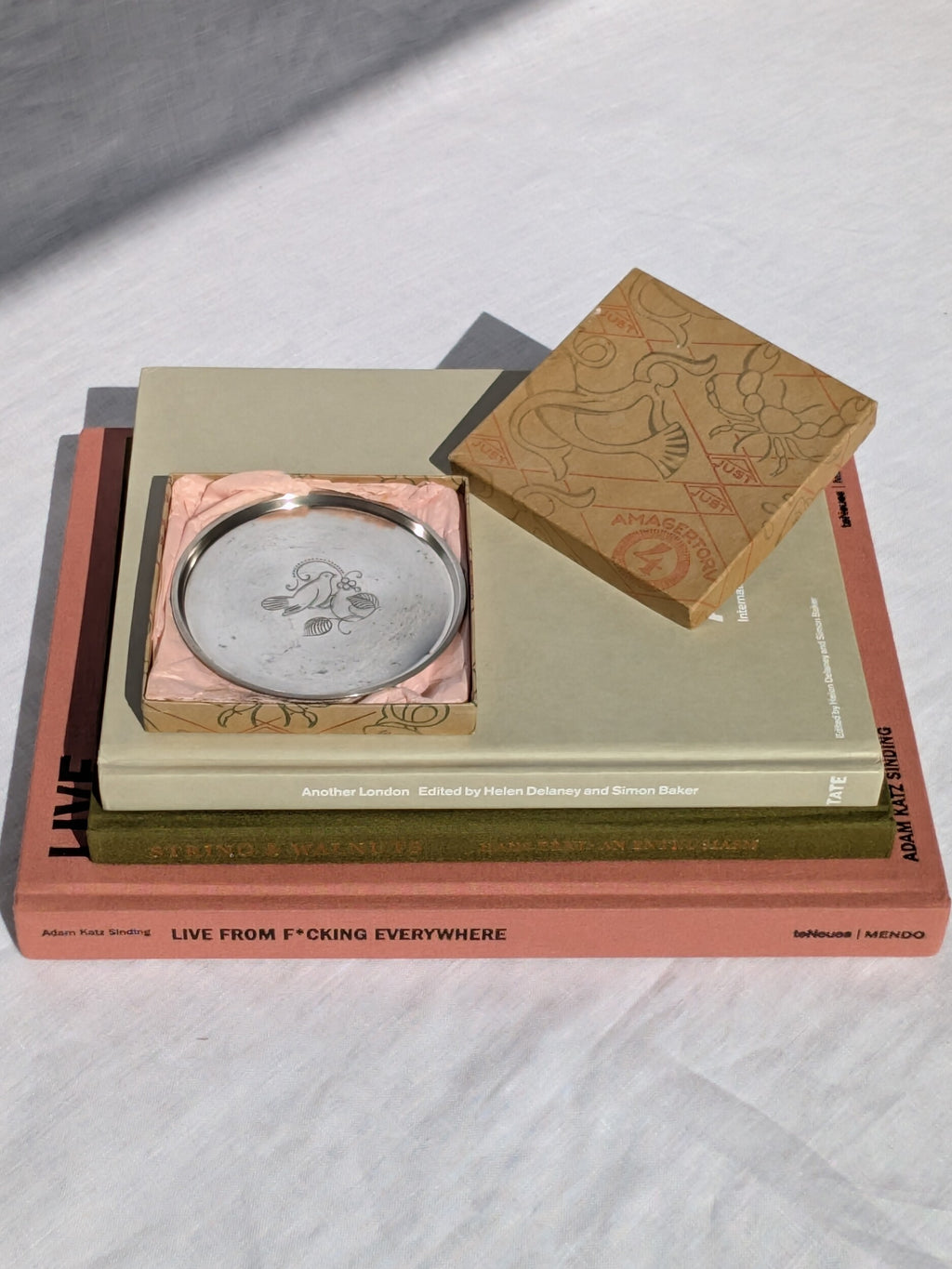 A 1930s vintage Just Andersen engraved pewter bottle coaster. An art deco circular silver coaster featuring a whimsical engraving of a dove on a branch in the centre. This collectable Danish mid century modern decor piece is styled on a stack of coffee table books inside the original Just Andersen gift box with baby pink tissue paper.