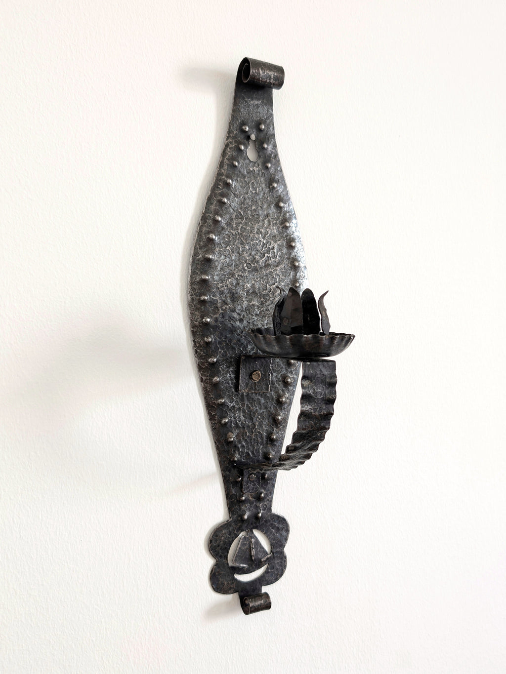 A vintage Spanish brutalist arts and crafts style candle wrought iron candle holder wall sconce on a white wall. This 1970s mid century modern Gothic inspired iron lighting piece is hand forged with a hammered texture, carved cut out elements, punched detailing and areas of organic patina. An interesting piece of iron metal wall art that will elevate modernist, rustic and antique homes.