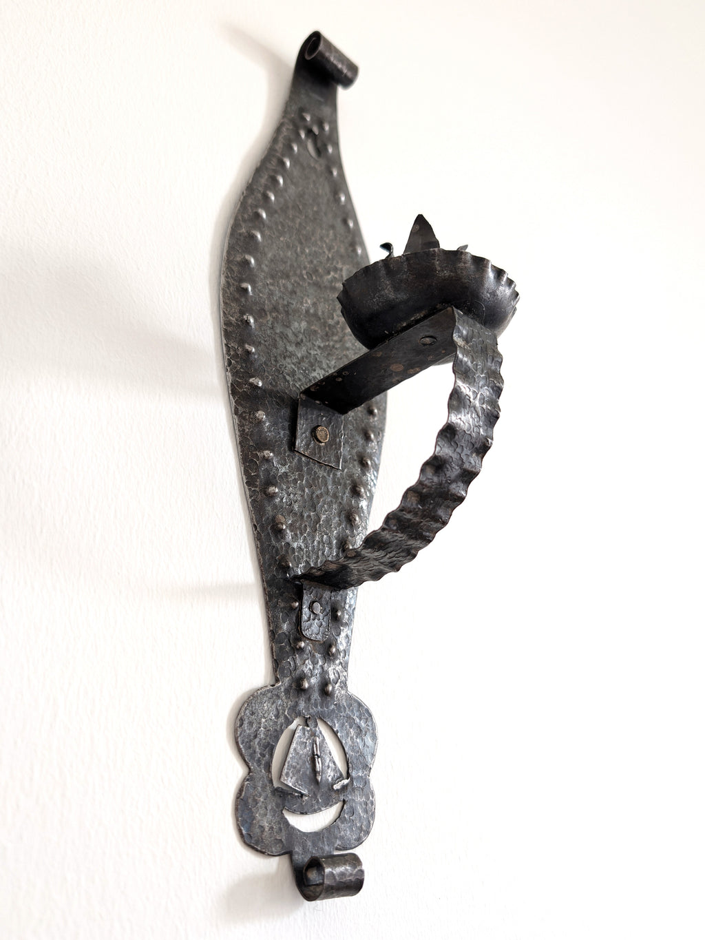 A vintage Spanish brutalist arts and crafts style candle wrought iron candle holder wall sconce on a white wall. This 1970s mid century modern Gothic inspired iron lighting piece is hand forged with a hammered texture, carved cut out elements, punched detailing and areas of organic patina. An interesting piece of iron metal wall art that will elevate modernist, rustic and antique homes.