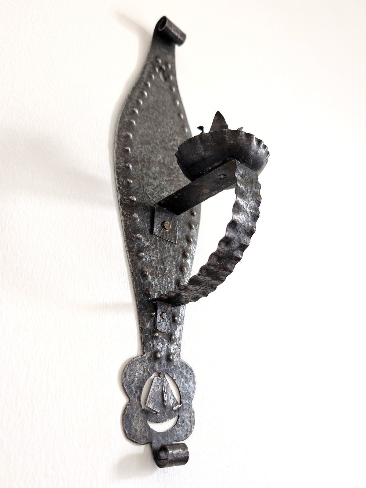A vintage Spanish brutalist arts and crafts style candle wrought iron candle holder wall sconce on a white wall. This 1970s mid century modern Gothic inspired iron lighting piece is hand forged with a hammered texture, carved cut out elements, punched detailing and areas of organic patina. An interesting piece of iron metal wall art that will elevate modernist, rustic and antique homes.