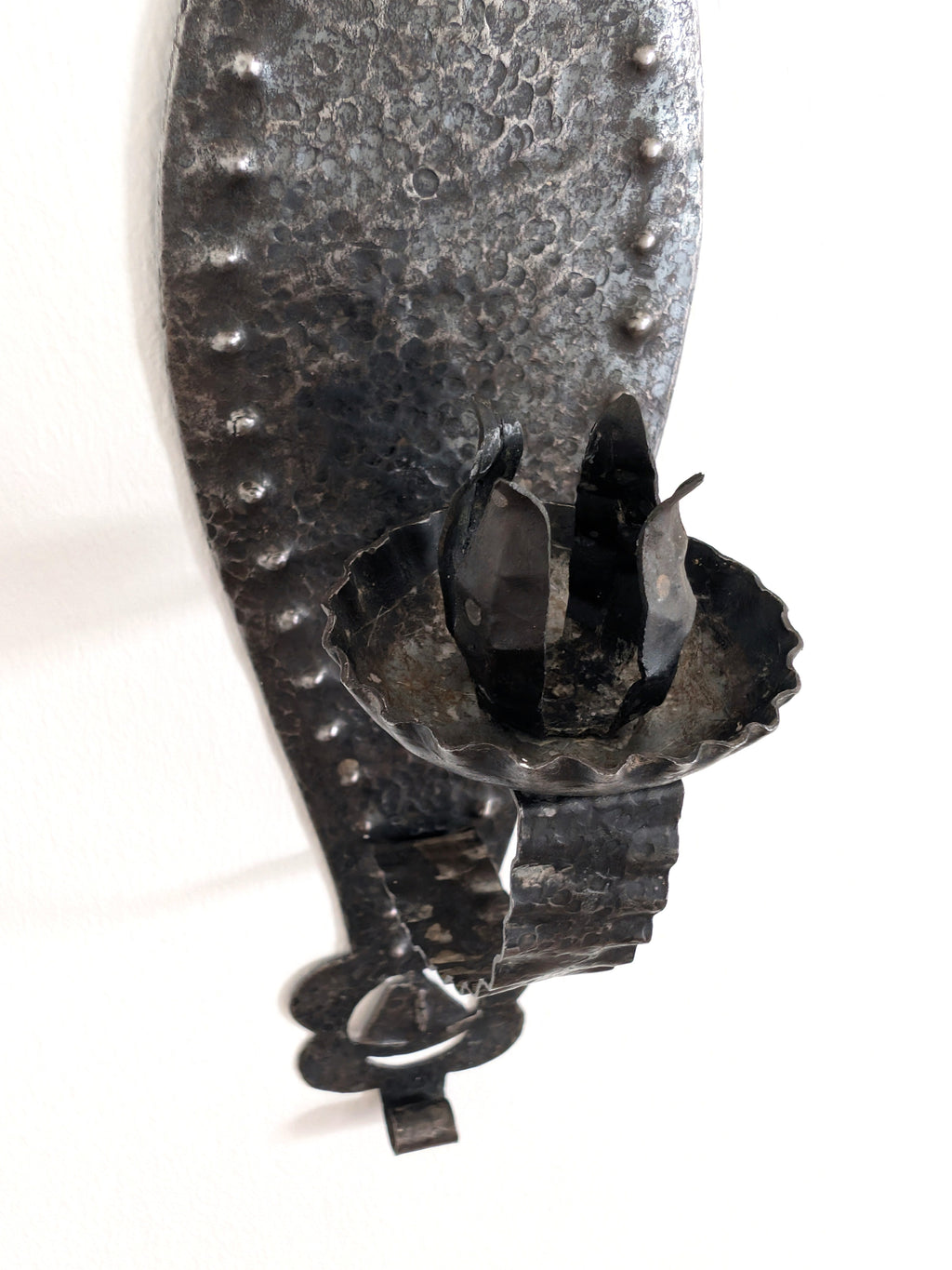 A close up of a vintage Spanish brutalist arts and crafts style candle wrought iron candle holder wall sconce on a white wall. This 1970s mid century modern Gothic inspired iron lighting piece is hand forged with a hammered texture, carved cut out elements, punched detailing and areas of organic patina. An interesting piece of iron metal wall art that will elevate modernist, rustic and antique homes.