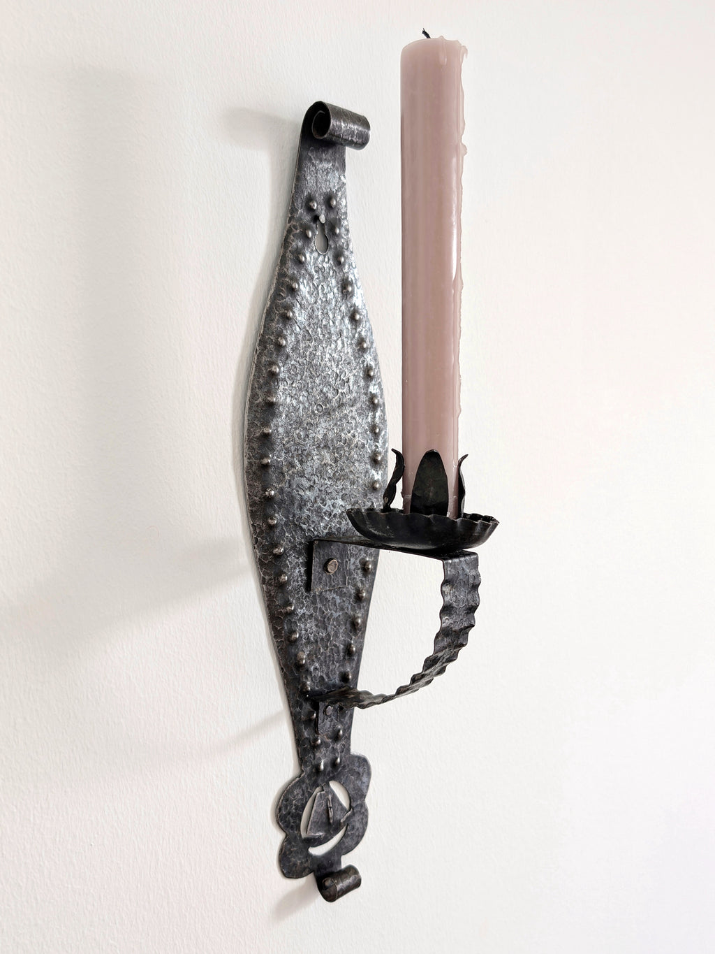 A vintage arts and crafts style candle wrought iron candle holder wall sconce on a white wall holding a slim pink candle. A mid century modern Gothic inspired iron lighting piece, hand forged with a hammered texture, carved cut outs, punched detailing and areas of organic patina, this metal wall art will elevate your home aesthetic with a timeless brutalist edge.