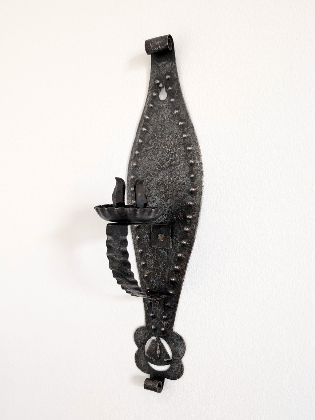 A vintage Spanish brutalist arts and crafts style candle wrought iron candle holder wall sconce on a white wall. This 1970s mid century modern Gothic inspired iron lighting piece is hand forged with a hammered texture, carved cut out elements, punched detailing and areas of organic patina. An interesting piece of iron metal wall art that will elevate modernist, rustic and antique homes.
