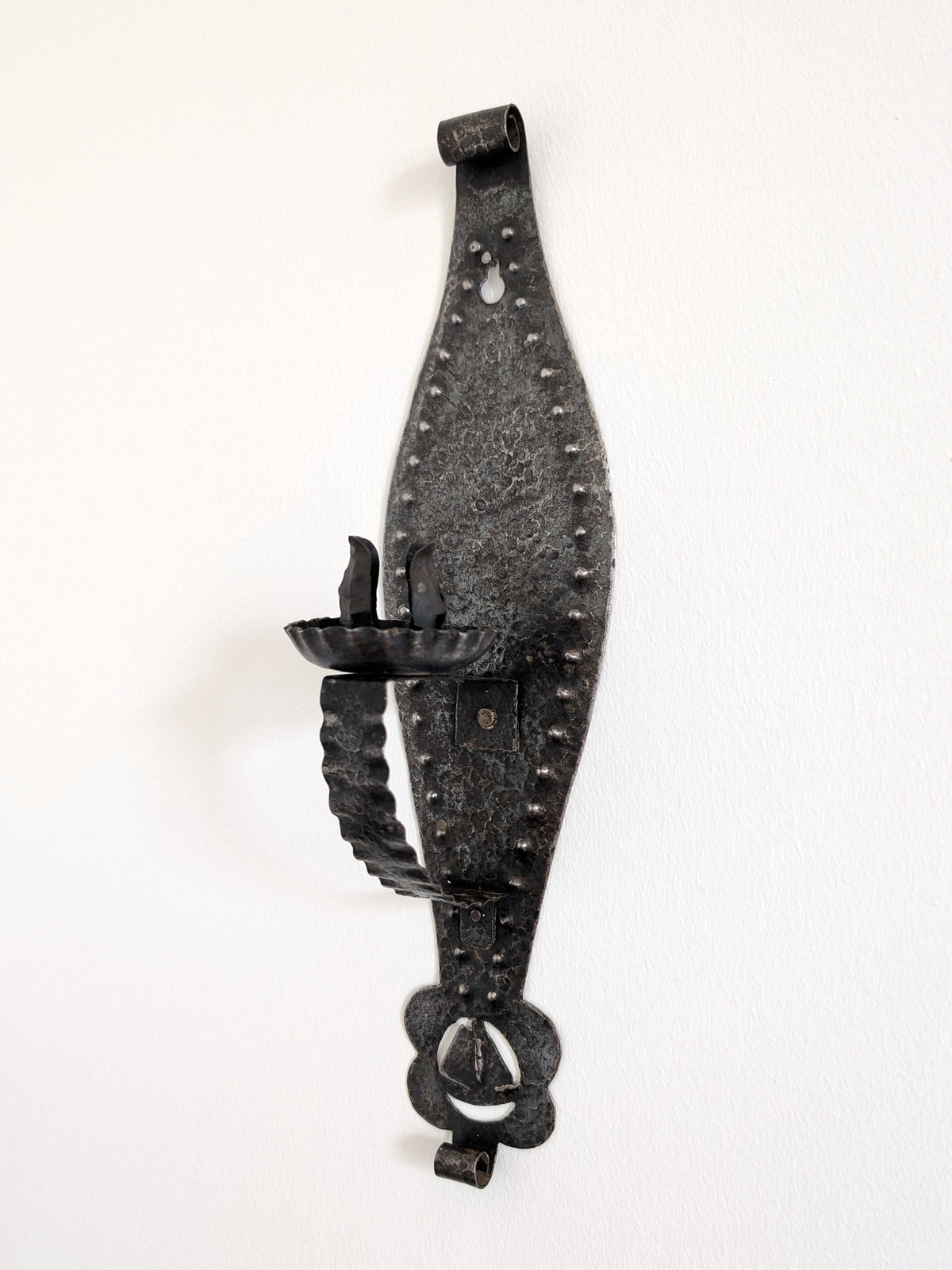 A vintage Spanish brutalist arts and crafts style candle wrought iron candle holder wall sconce on a white wall. This 1970s mid century modern Gothic inspired iron lighting piece is hand forged with a hammered texture, carved cut out elements, punched detailing and areas of organic patina. An interesting piece of iron metal wall art that will elevate modernist, rustic and antique homes.