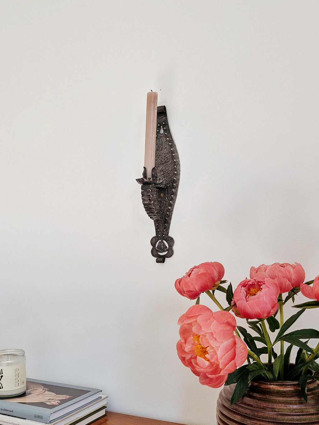 A vintage arts and crafts movement black and dark silver wrought iron candle holder wall sconce on a white wall behind an art deco terracotta ceramic vase containing pink peonies and a stack of books with a cream candle on top. A mid century modern Gothic style metal wall lighting piece, hand forged with a hammered texture, carved cut outs, punched detailing and areas of organic rustic patina, this antique inspired metal wall decor will lend a timeless brutalist design element to your home.