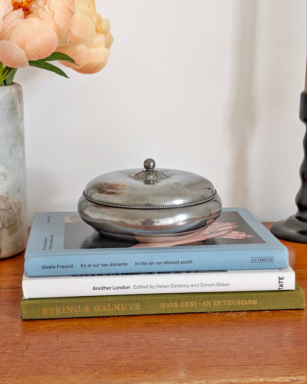 A 1930s vintage Just Andersen pewter lidded trinket box with a floral handle and beaded detailing around the lid edge. This Danish art deco trinket holder is styled on a stack of coffee table books next to a minimal grey marble vase filled with peach peonies and a black candle holder in front of a white wall. This rare mid century modern design would make a stylish jewellery holder in modernist, minimalist, vintage modern, Hollywood Regency or antique baroque homes.