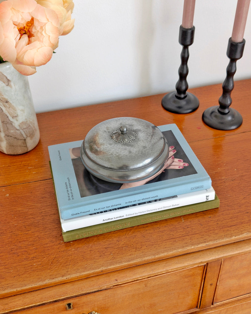 A 1930s vintage Just Andersen pewter lidded trinket box with a floral handle and beaded detailing around the lid edge. This Danish art deco trinket holder is styled on a stack of coffee table books next to a minimal grey marble vase filled with peach peonies in front of a white wall. This rare mid century modern design would make a stylish jewellery holder in modernist, minimalist, vintage modern, Hollywood Regency or antique baroque home interior schemes.