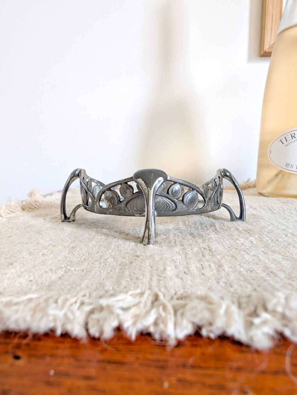 A close up of an antique art nouveau pewter bottle holder by Archibald Knox for Liberty & Co next to a glass of wine on a green marble coaster with a white wall in the background. Dated 1905, this rare silver wine bottle holder features an aesthetic movement style hand carved floral cut out design and is raised by four curved lug feet.