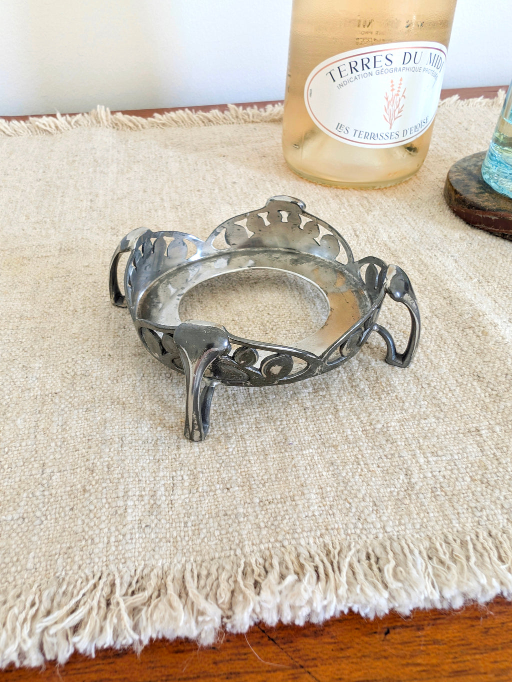 A close up of an antique art nouveau pewter bottle holder by Archibald Knox for Liberty & Co next to a glass of wine on a green marble coaster with a white wall in the background, shot from above to show the inside. Dated 1905, this rare silver wine bottle holder features an aesthetic movement style hand carved floral cut out design and is raised by four curved lug feet.