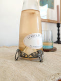 A close up of an antique art nouveau pewter bottle holder by Archibald Knox for Liberty & Co, London holding a bottle of rosé wine on next to a glass of wine on a green marble coaster with a black wooden candleholder and a vintage original multicoloured painting on a white wall in the background. Dated 1905, this rare silver wine bottle holder features an aesthetic movement style hand carved floral cut out design and is raised by four curved lug feet.