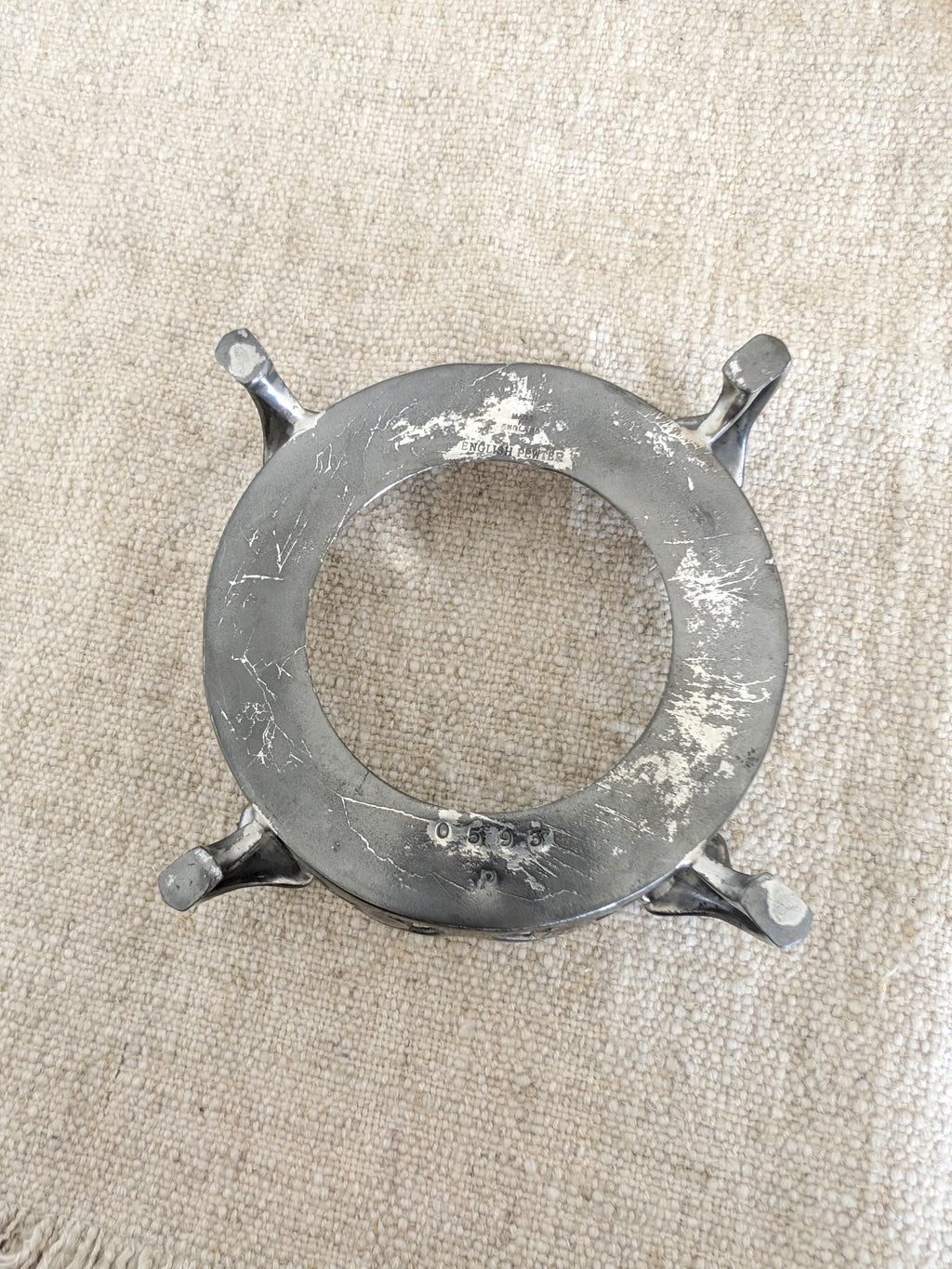 A close up of the base of an antique art nouveau pewter bottle holder by Archibald Knox for Liberty & Co, on a  rustic linen surface showing the makers signature 'Made In England', 'English Pewter' and the model number '0593' engraved to provide the authenticity of the piece. Dated 1905, this rare silver wine bottle holder features an aesthetic movement style floral cut out design, areas of pewter patina and is raised by four curved lug feet.