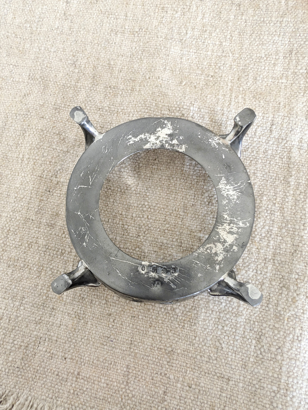 A close up of the base of an antique art nouveau pewter bottle holder by Archibald Knox for Liberty & Co, on a  rustic linen surface showing the makers signature 'Made In England', 'English Pewter' and the model number '0593' engraved to provide the authenticity of the piece. Dated 1905, this rare silver wine bottle holder features an aesthetic movement style floral cut out design, areas of pewter patina and is raised by four curved lug feet.