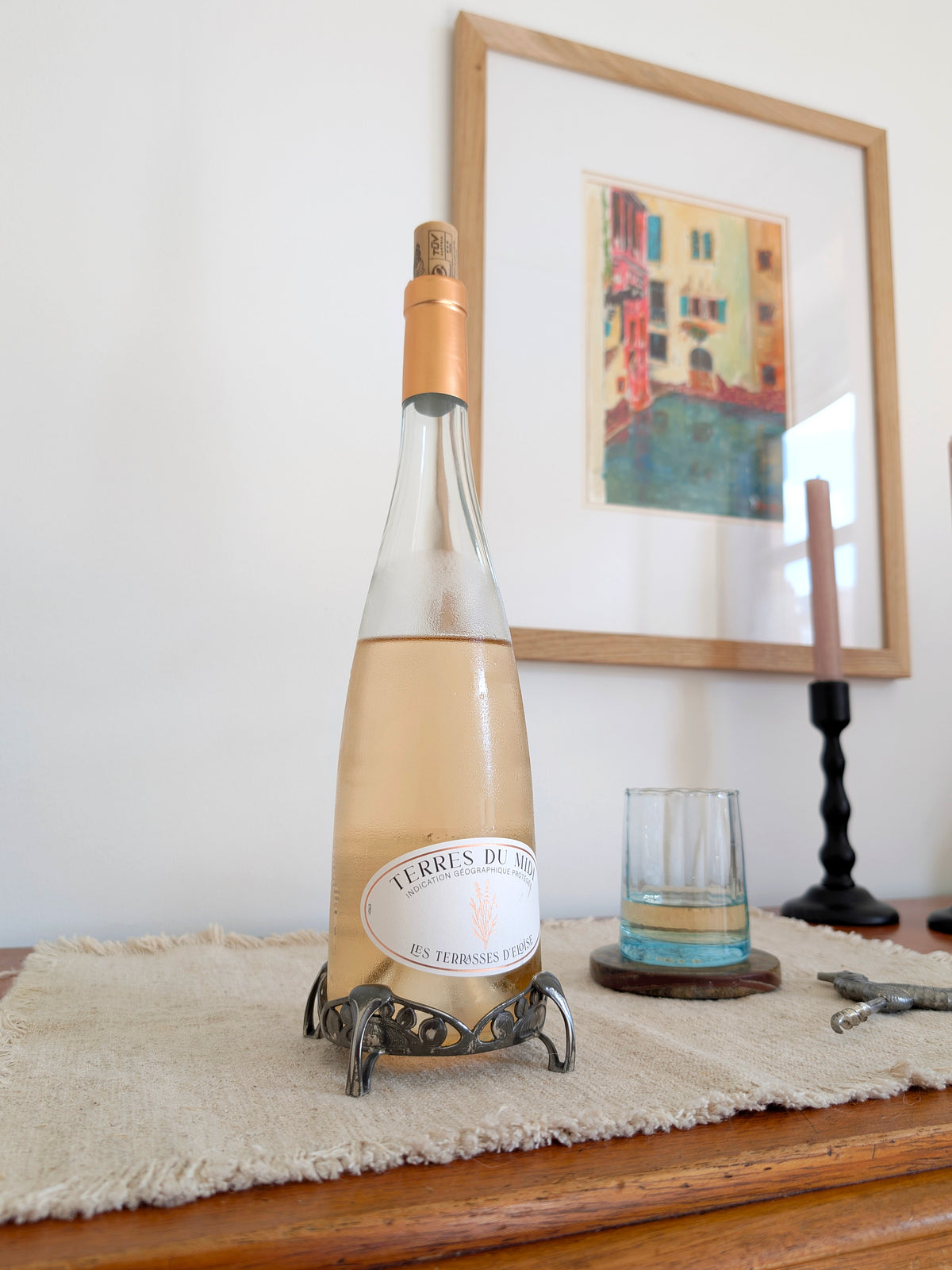 An antique art nouveau pewter bottle holder by Archibald Knox for Liberty & Co holding a bottle of rosé wine on next to a glass of wine on a green marble coaster, a black wooden candleholder holding, an art deco pewter Just Andersen bottle opener and a vintage multicoloured painting on a white wall in the background. Dated 1905, this rare silver wine bottle holder features an aesthetic movement style hand carved floral cut out design and is raised by four curved lug feet.