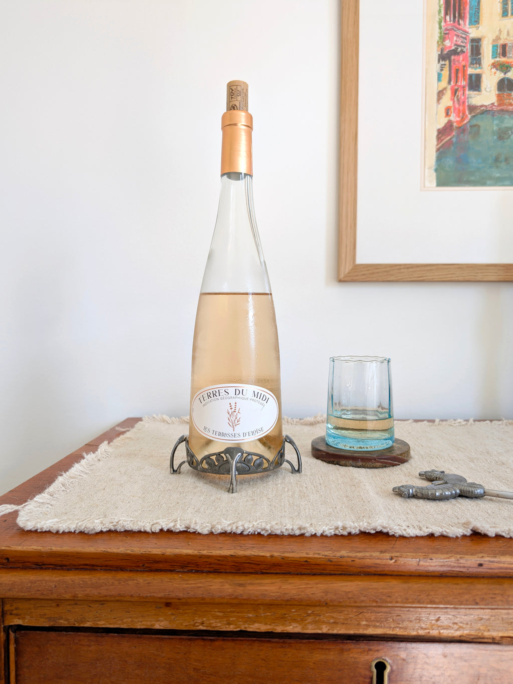 An antique art nouveau pewter bottle holder by Archibald Knox for Liberty & Co holding a bottle of rosé wine on next to a glass of wine on a green marble coaster, an art deco pewter Just Andersen bottle opener and a vintage multicoloured painting on a white wall in the background. Dated 1905, this rare silver wine bottle holder features an aesthetic movement style hand carved floral cut out design and is raised by four curved lug feet.