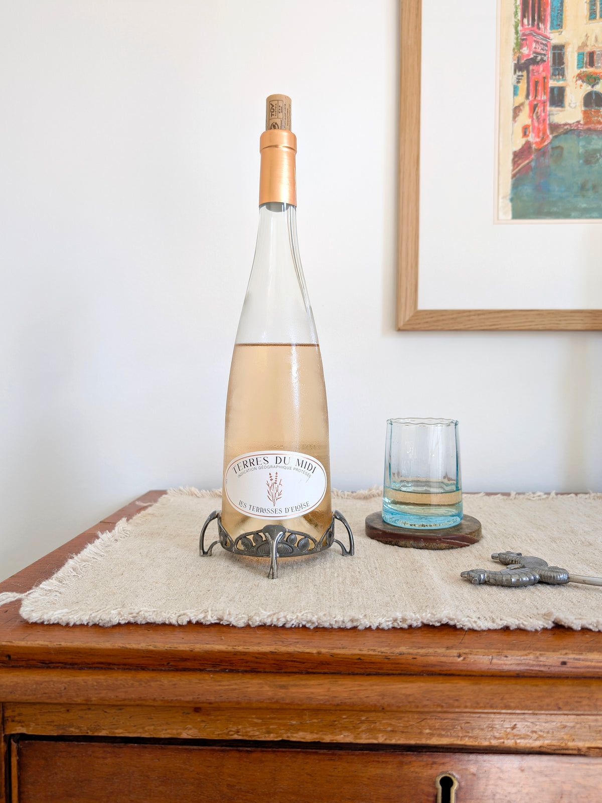An antique art nouveau pewter bottle holder by Archibald Knox for Liberty & Co holding a bottle of rosé wine on next to a glass of wine on a green marble coaster, an art deco pewter Just Andersen bottle opener and a vintage multicoloured painting on a white wall in the background. Dated 1905, this rare silver wine bottle holder features an aesthetic movement style hand carved floral cut out design and is raised by four curved lug feet.
