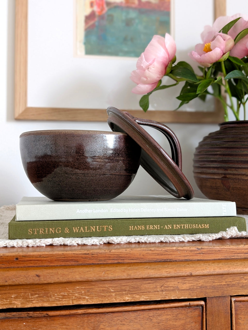 A vintage 1960s tenmoku glaze ceramic lidded pot by Henry Hammond styled on a stack of coffee table books with an art deco ridged vase with pink peonies and a colourful watercolour painting in the background. This piece of mid century modern studio pottery earthenware features an iridescent terracotta and black glaze and an off white speckled inner glaze, this beautifully crafted studio pottery bowl would blend perfectly with rustic cottage farmhouse, modernist or minimalist homes.