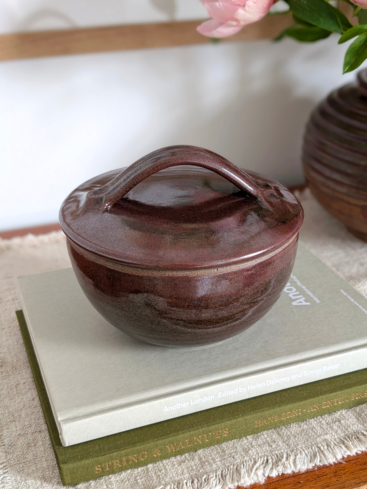 A vintage 1960s tenmoku glaze ceramic lidded pot by Henry Hammond styled on a stack of coffee table books with an art deco ridged vase with pink peonies and a colourful watercolour painting in the background. This piece of mid century modern studio pottery earthenware features an iridescent terracotta and black glaze and an off white speckled inner glaze, this beautifully crafted studio pottery bowl would blend perfectly with rustic cottage farmhouse, modernist or minimalist homes.