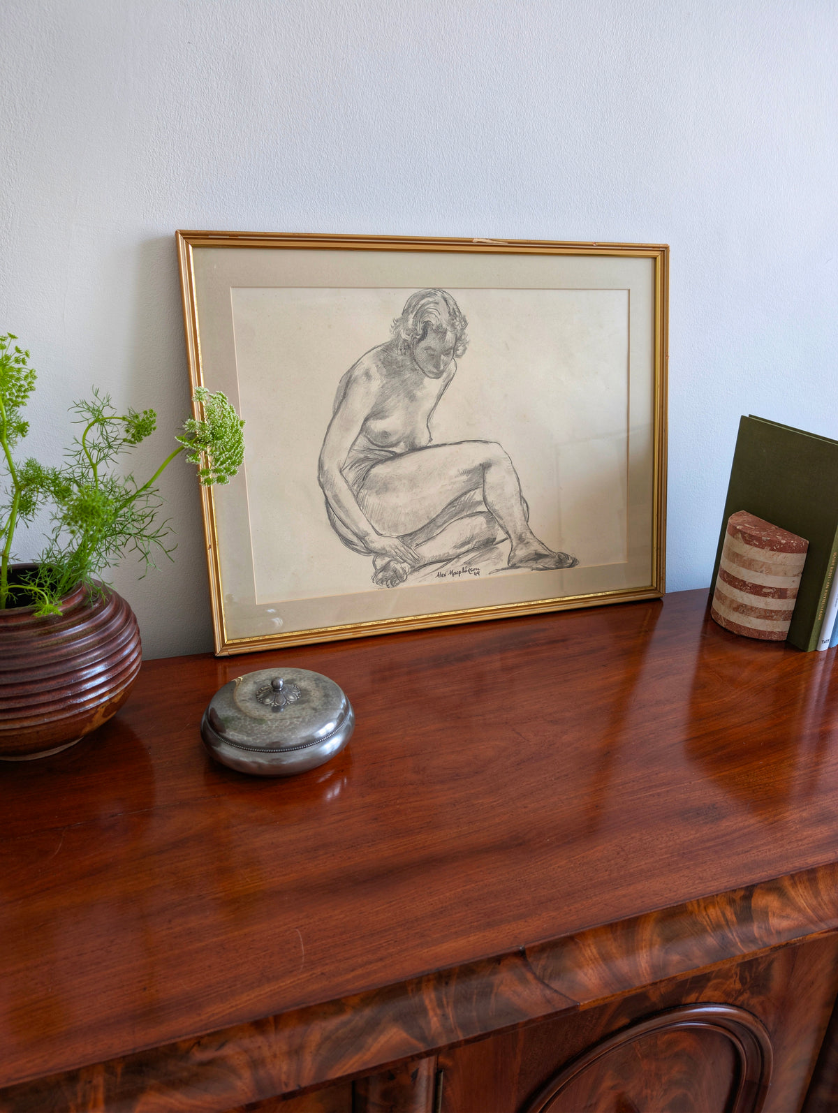 Vintage original charcoal female nude study portrait by Alexander MacPherson on an antique side board next to a brutalist vase, an art deco pewter pot and mid century modern striped bookends in front of a white wall. This monochrome, minimalist artwork of a serene seated model is signed and dated by the artist and has a gold wooden frame. The work holds universal appeal, blending seamlessly within rustic, farmhouse, cottage or heritage home interior schemes.
