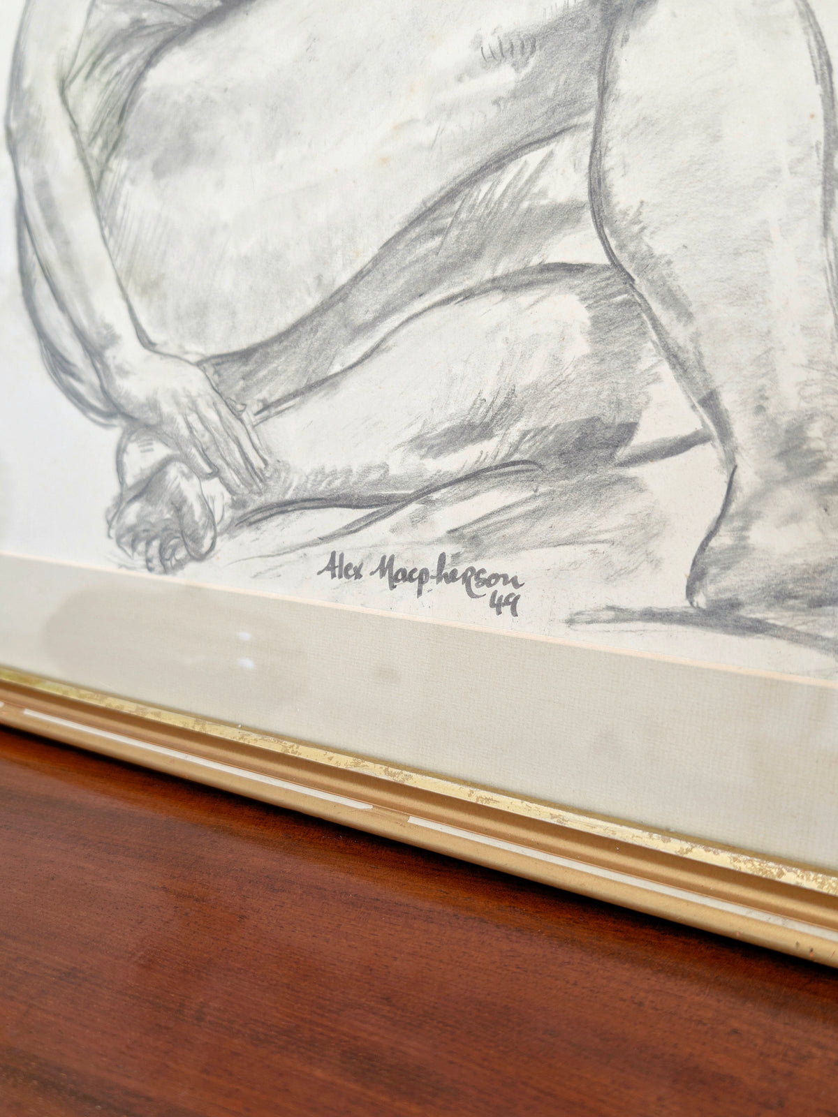 A close up of the artist' signature on a vintage original charcoal female nude study portrait by Alexander MacPherson on an antique side board next to a brutalist vase, an art deco pewter pot and mid century modern striped bookends in front of a white wall. This monochrome, minimalist artwork of a serene seated model is signed and dated by the artist and has a gold wooden frame. The work holds universal appeal, blending seamlessly within rustic, farmhouse, cottage or heritage home interior schemes.