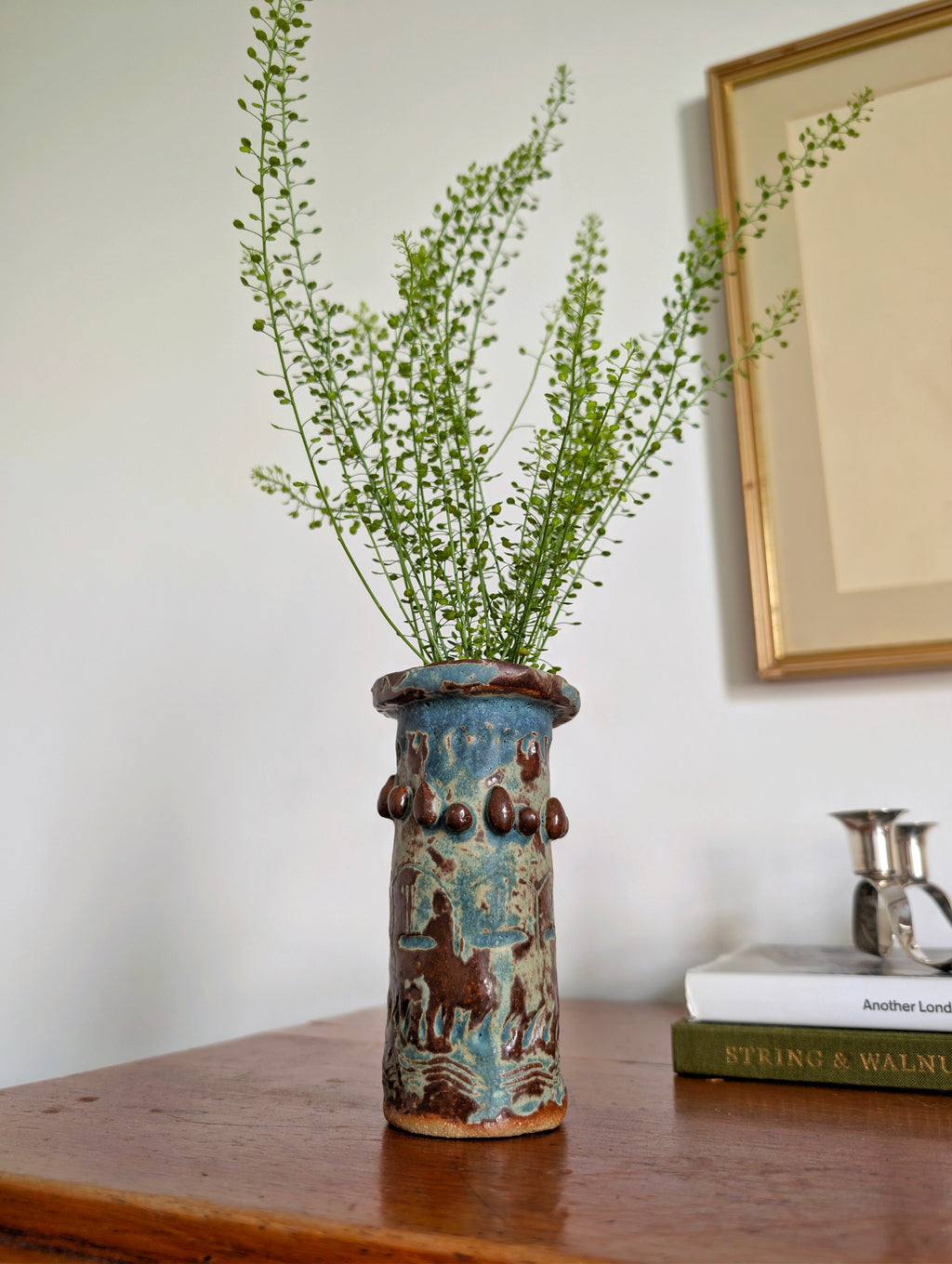 A vintage brutalist ceramic vase with a matte blue, green and brown glaze next to an antique silver candleholder on a wood sideboard in front a white wall with a vintage artwork hanging on it. This 1970s primitive pottery piece features irregular design details such as abstract engravings and textural surface elements which make this studio pottery object ideal for those looking to add an interesting visual statement to mid century, modernist, rustic, retro, industrial or eclectic home interiors.