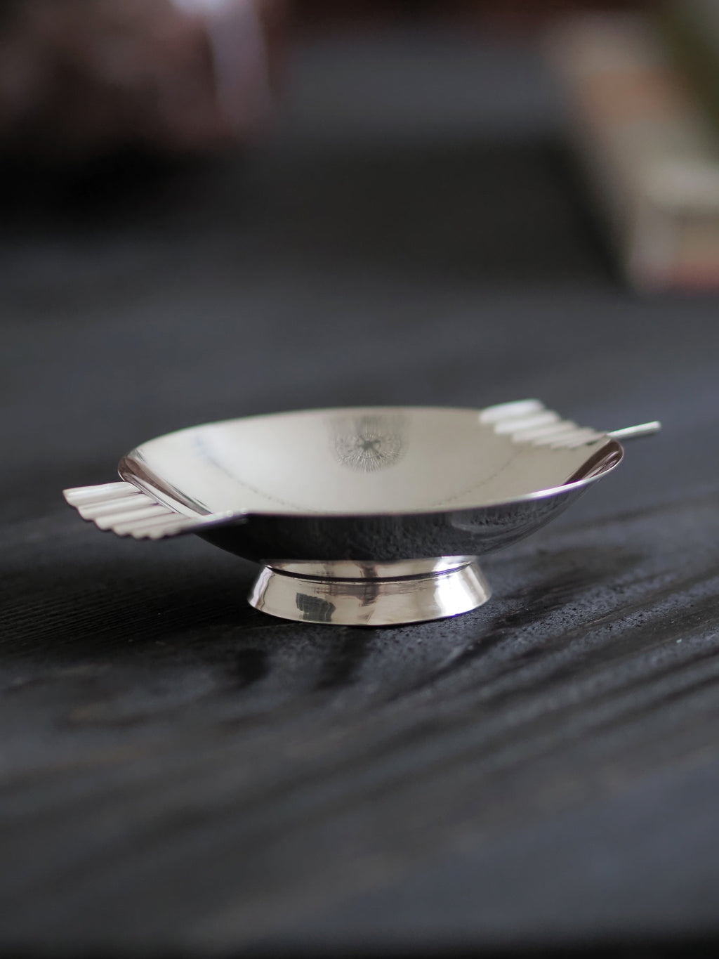The side of a vintage modernist silver ring dish by James Dixon & Sons styled on black wood surface. Crafted from silver plate, this petite circular footed trinket dish has geometric ridged lugs and would compliment postmodern, Swedish grace, minimalist, mid century modern, Bauhaus, art deco and Hollywood Regency décor schemes. Sold by Vintrovia, vintage home décor store UK.
