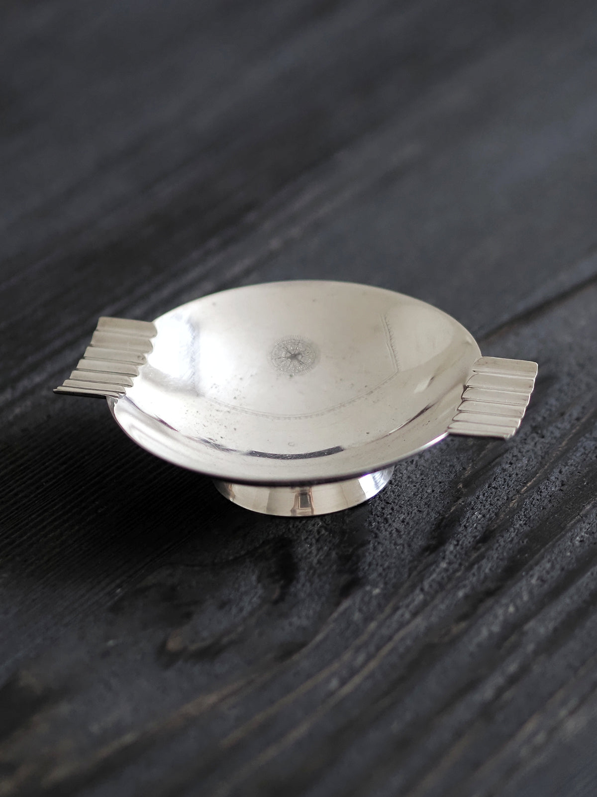 A vintage modernist silver ring dish by James Dixon & Sons styled on black wood surface. Crafted from silver plate, this petite circular footed trinket dish has geometric ridged lugs and would compliment postmodern, Swedish grace, minimalist, mid century modern, Bauhaus, art deco and Hollywood Regency décor schemes. Sold by Vintrovia, vintage home décor store UK.