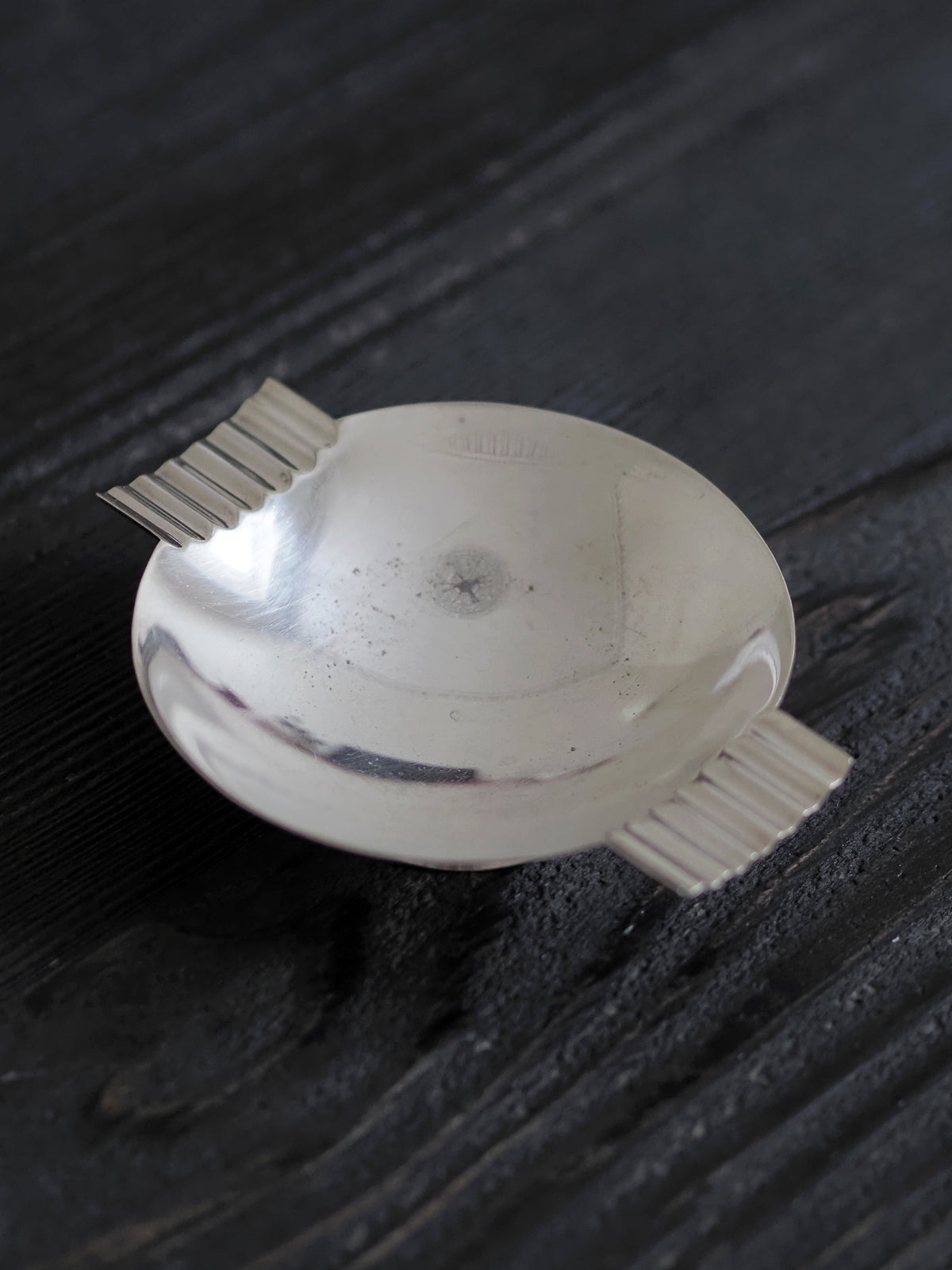 A close up of a 1920s vintage modernist silver ring dish by James Dixon & Sons styled on black wood surface. Crafted from silver plate, this petite circular footed trinket dish has geometric ridged lugs and would compliment postmodern, Swedish grace, minimalist, mid century modern, Bauhaus, art deco and Hollywood Regency décor schemes. Sold by Vintrovia, vintage home décor store UK.
