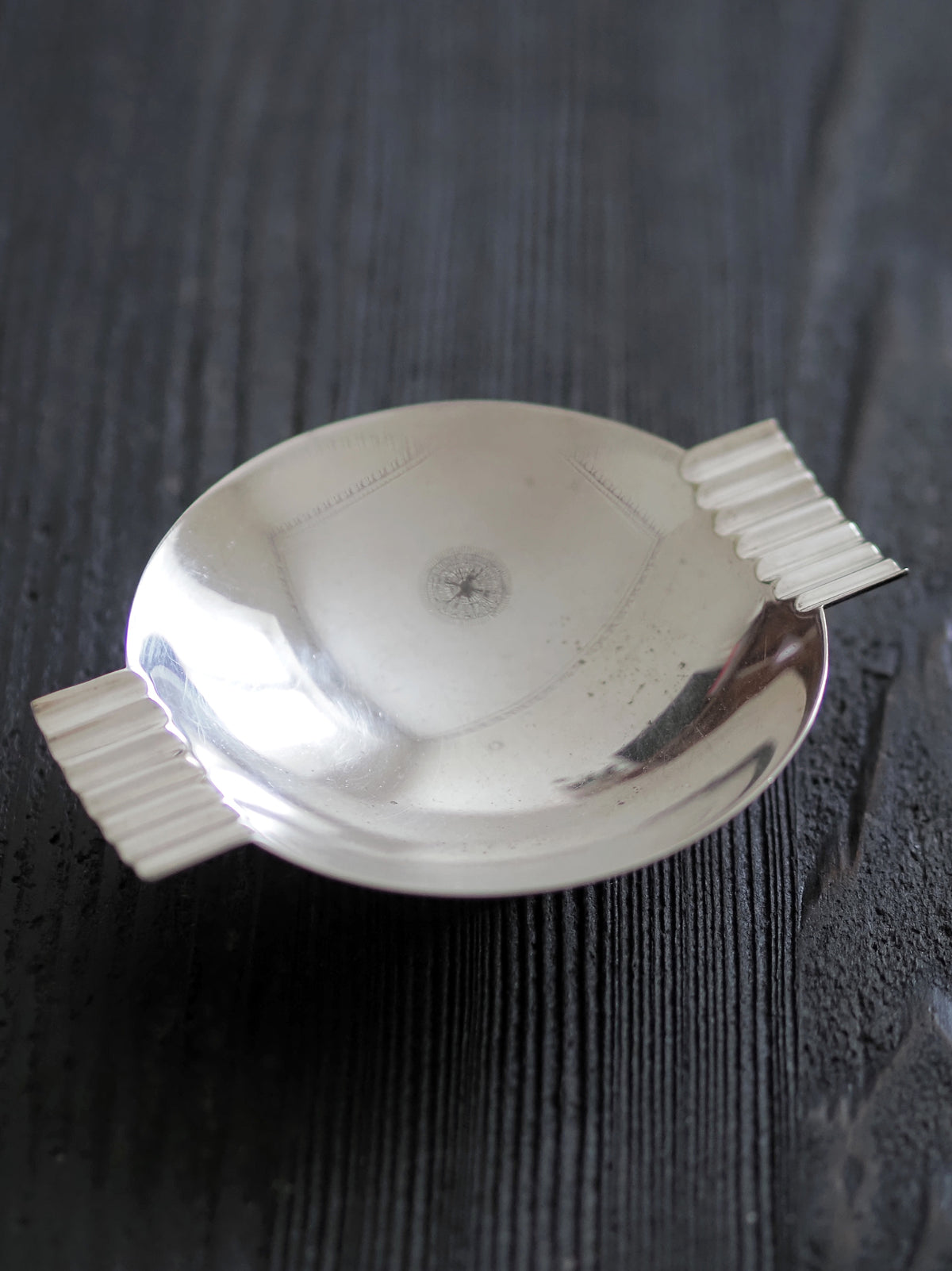 A vintage modernist silver ring dish by James Dixon & Sons styled on black wood surface. Crafted from silver plate, this petite circular footed trinket dish has geometric ridged lugs and would compliment postmodern, Swedish grace, minimalist, mid century modern, Bauhaus, art deco and Hollywood Regency décor schemes. Sold by Vintrovia, vintage home décor store UK.