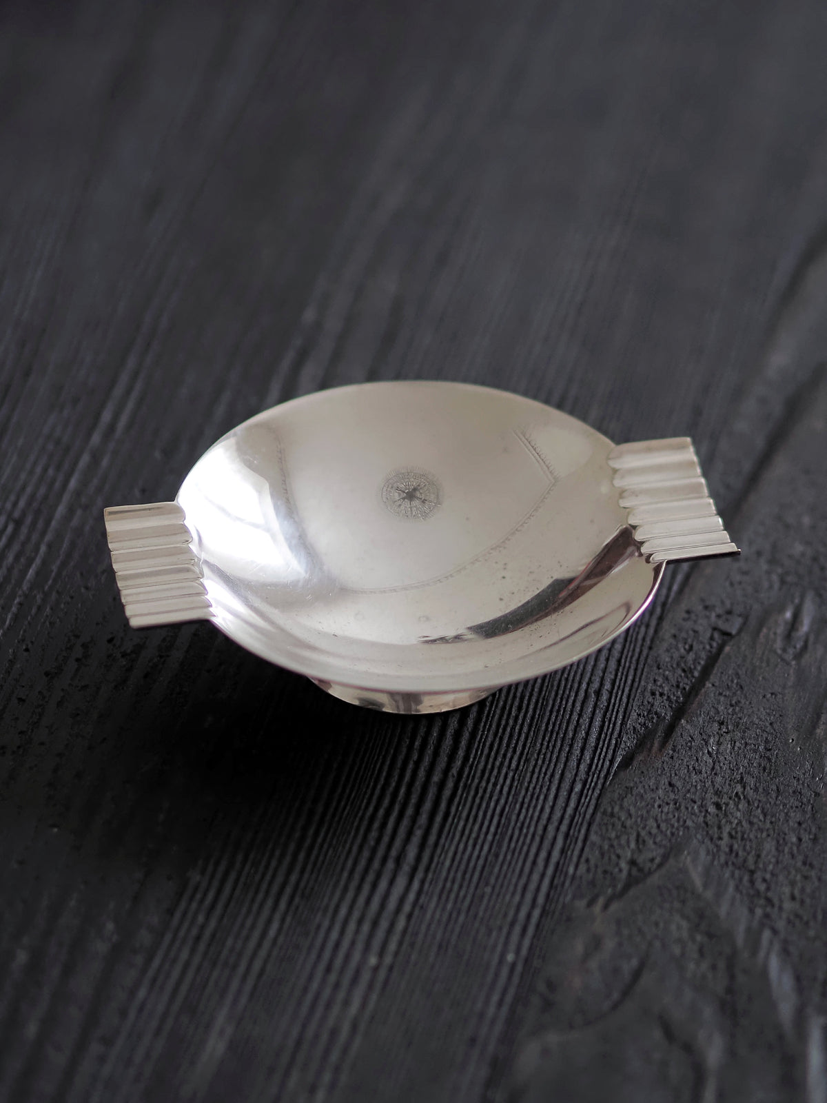 A close up of a 1920s vintage modernist silver ring dish by James Dixon & Sons styled on black wood surface. Crafted from silver plate, this petite circular footed trinket dish has geometric ridged lugs and would compliment postmodern, Swedish grace, minimalist, mid century modern, Bauhaus, art deco and Hollywood Regency décor schemes. Sold by Vintrovia, vintage home décor store UK.