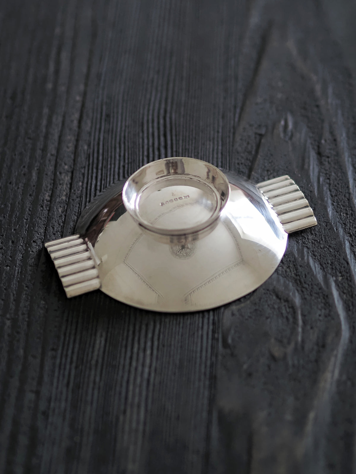 An upturned 1920s vintage modernist silver ring dish by James Dixon & Sons styled on black wood surface. Crafted from silver plate, this petite circular footed trinket dish has geometric ridged lugs and would compliment postmodern, Swedish grace, minimalist, mid century modern, Bauhaus, art deco and Hollywood Regency décor schemes. Sold by Vintrovia, vintage home décor store UK.