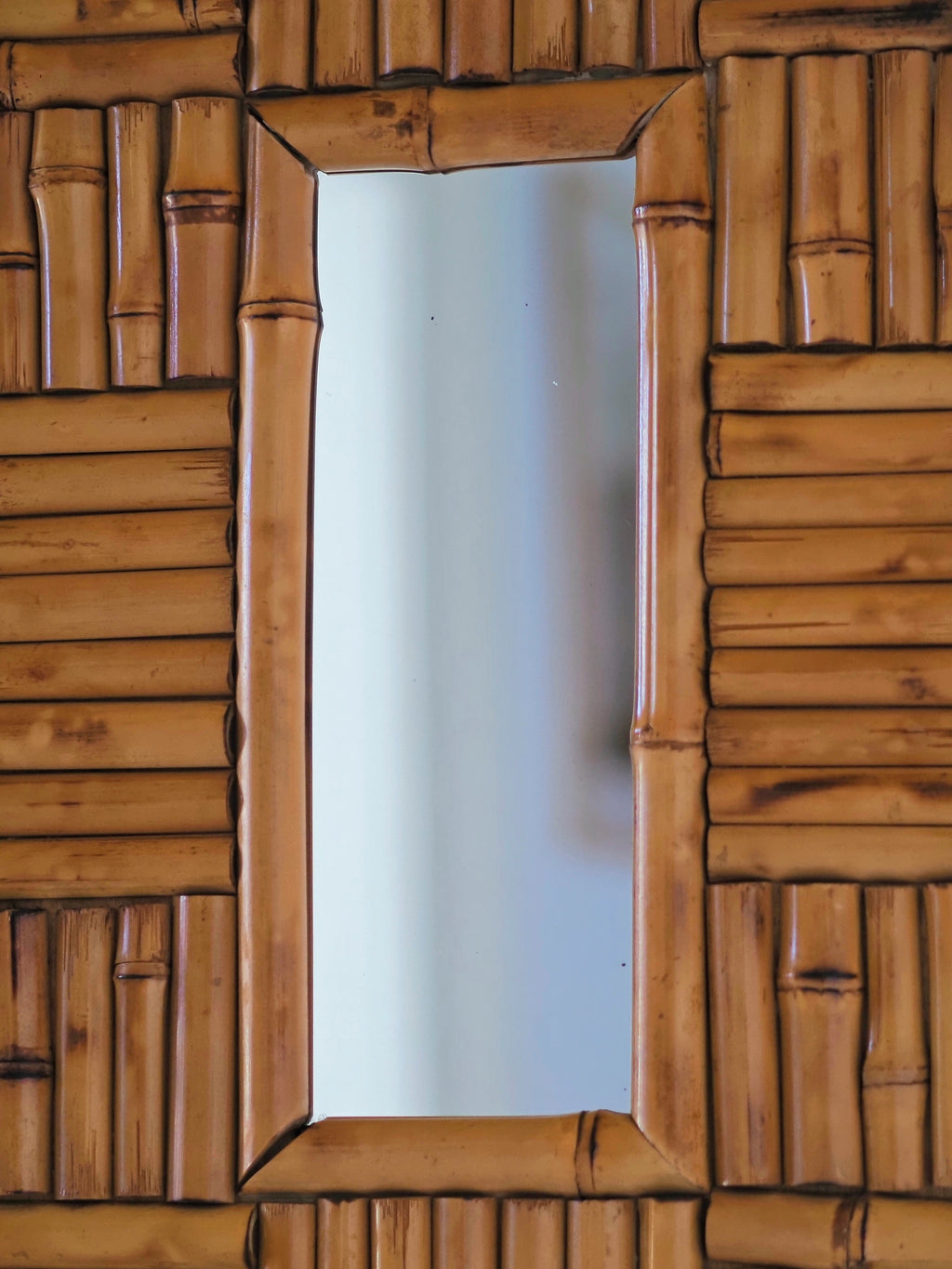 A close up of the centre of a vintage 1970s wall mounted bamboo mirror by UK modern vintage home décor store Vintrovia on a yellow wall. This mid century modern mirror is hand crafted using individual honey-toned rattan pieces to form a geometric check design that creates an interesting, modernist aesthetic. A versatile retro wall décor piece that would elevate boho, eclectic, coastal and rustic home interiors.
