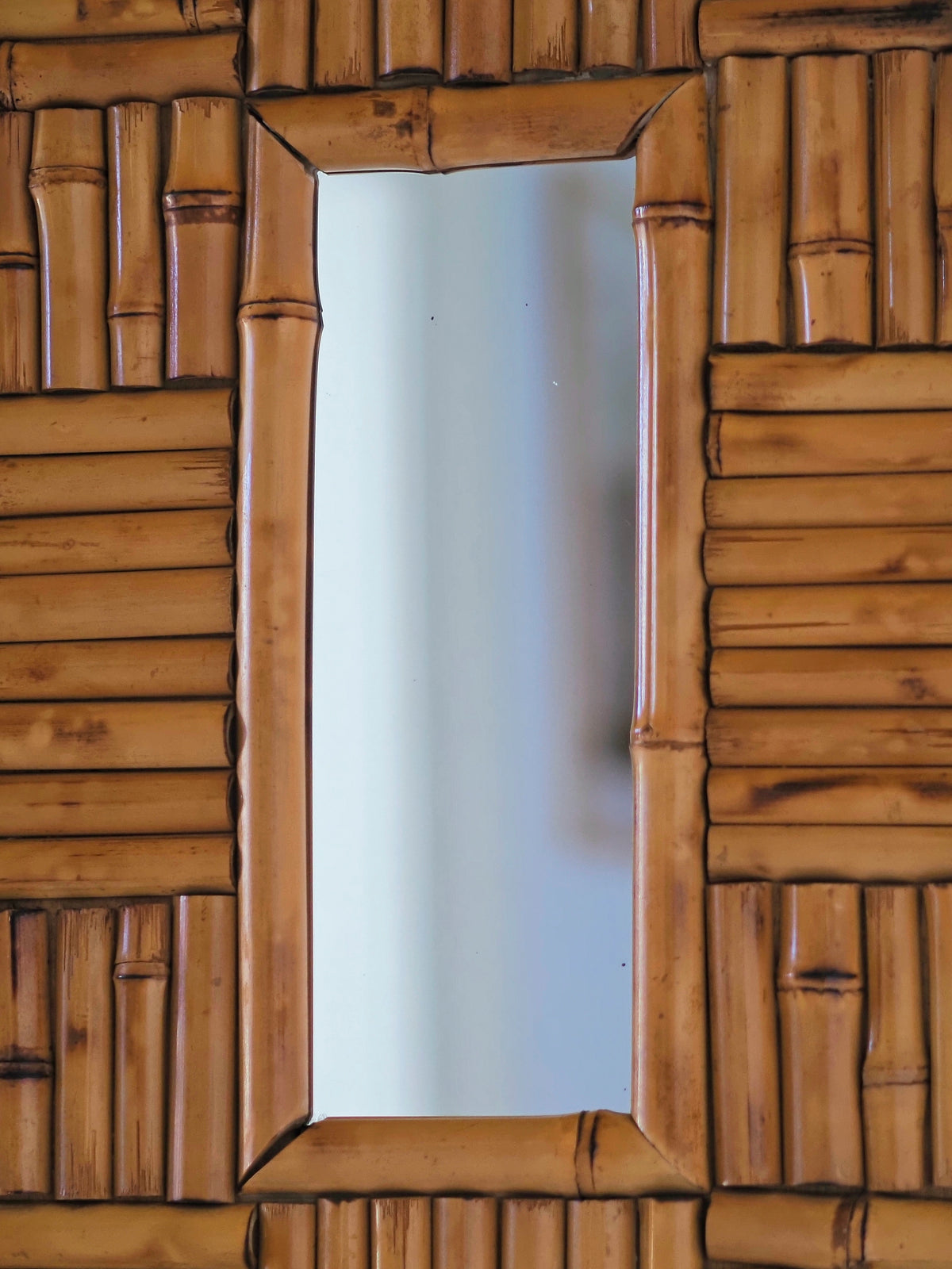 A close up of the centre of a vintage 1970s wall mounted bamboo mirror by UK modern vintage home décor store Vintrovia on a yellow wall. This mid century modern mirror is hand crafted using individual honey-toned rattan pieces to form a geometric check design that creates an interesting, modernist aesthetic. A versatile retro wall décor piece that would elevate boho, eclectic, coastal and rustic home interiors.