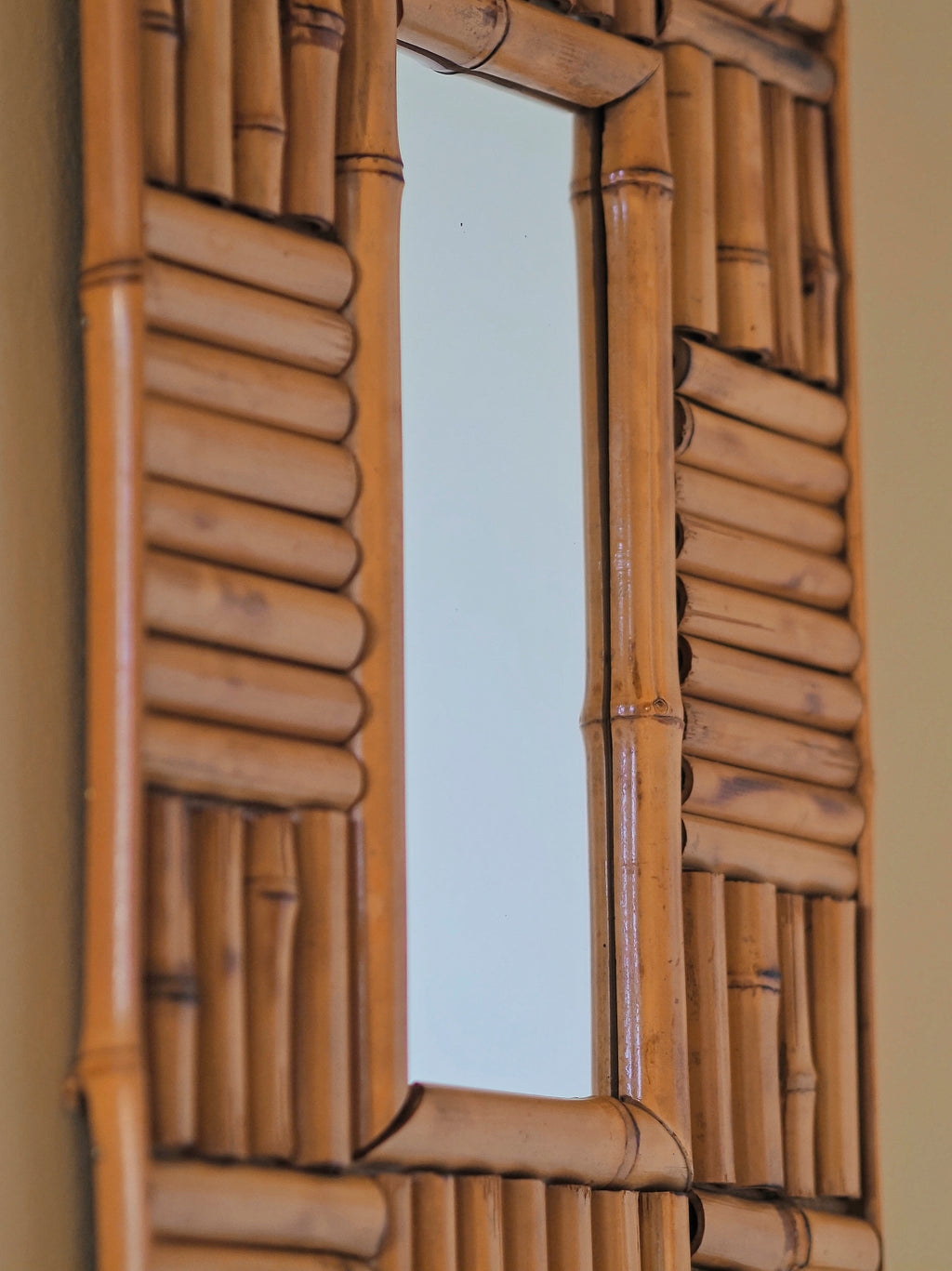 The side of a vintage 1970s wall mounted bamboo mirror by UK modern vintage home décor store Vintrovia on a yellow wall. This mid century modern mirror is hand crafted using individual honey-toned rattan pieces to form a geometric check design that creates an interesting, modernist aesthetic. A versatile retro wall décor piece that would elevate boho, eclectic, coastal and rustic home interiors.
