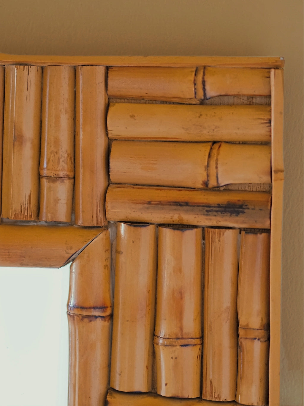 A close up of a vintage 1970s wall mounted bamboo mirror by UK modern vintage home décor store Vintrovia on a yellow wall. This mid century modern mirror is hand crafted using individual honey-toned rattan pieces to form a geometric check design that creates an interesting, modernist aesthetic. A versatile retro wall décor piece that would elevate boho, eclectic, coastal and rustic home interiors.