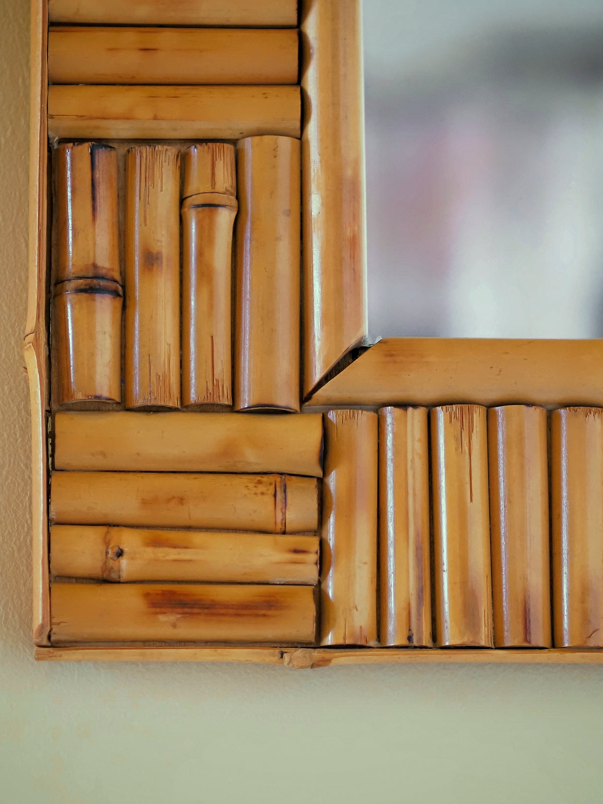 The corner of a vintage 1970s wall mounted bamboo mirror by UK modern vintage home décor store Vintrovia on a yellow wall. This mid century modern mirror is hand crafted using individual honey-toned rattan pieces to form a geometric check design that creates an interesting, modernist aesthetic. A versatile retro wall décor piece that would elevate boho, eclectic, coastal and rustic home interiors.