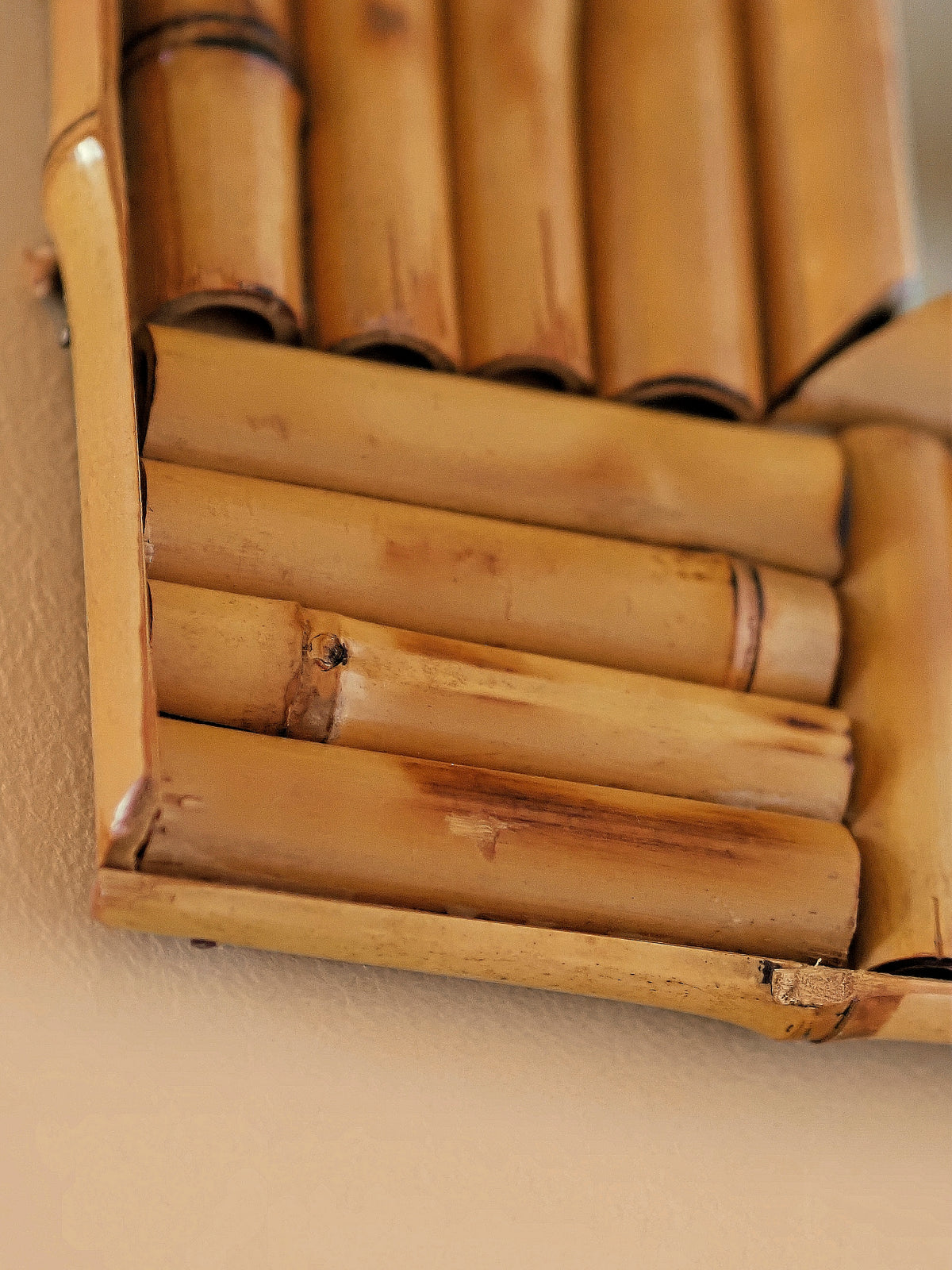A close up of a vintage 1970s wall mounted bamboo mirror by UK modern vintage home décor store Vintrovia on a yellow wall. This mid century modern mirror is hand crafted using individual honey-toned rattan pieces to form a geometric check design that creates an interesting, modernist aesthetic. A versatile retro wall décor piece that would elevate boho, eclectic, coastal and rustic home interiors.