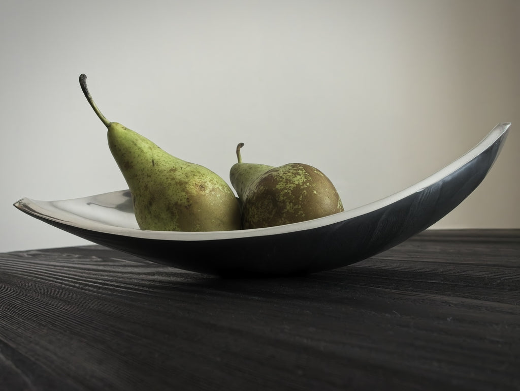 Two green pears in a silver mid century modern vintage sculptural bowl on a black wooden surface with a neutral background.