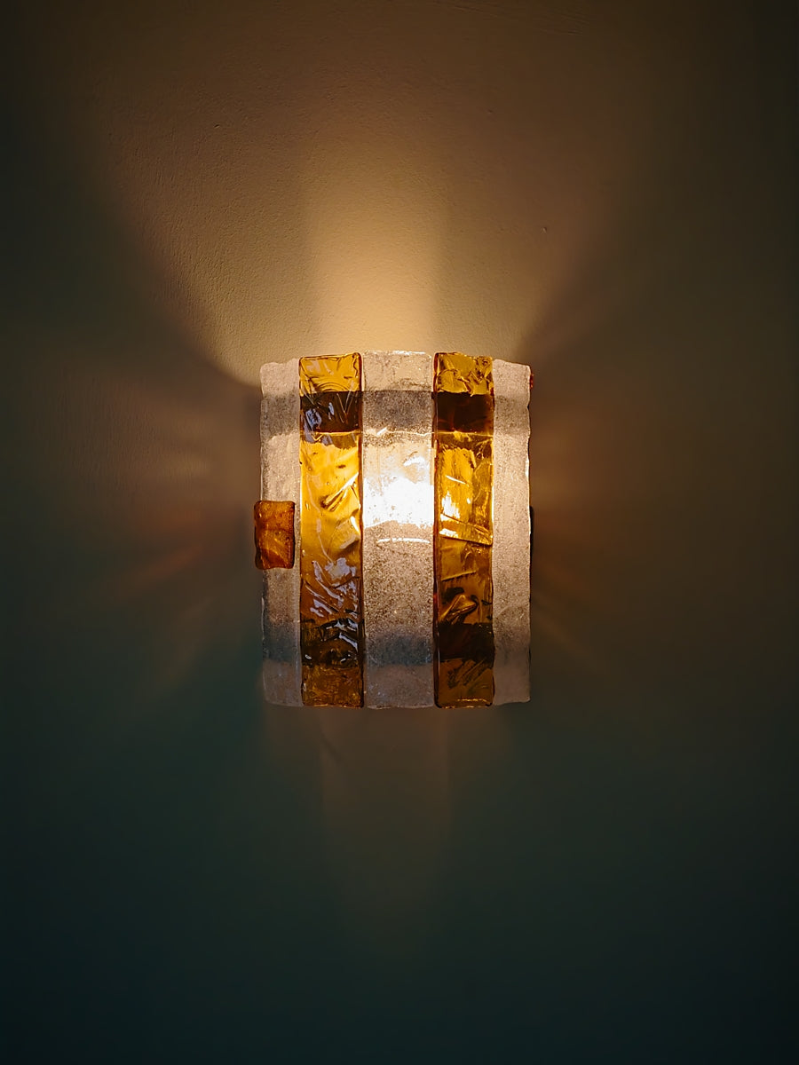 Wall-mounted light fixture with striped amber and clear glass panels on a dark background with light beams radiating outwards