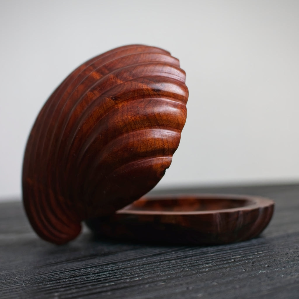 A close up of a small mid century modern 1960s vintage wooden shell shaped trinket box with a carved, ridged lid on a dark surface.