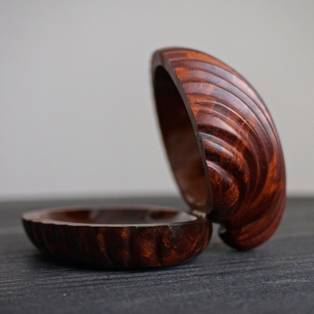 A close up of a vintage mid century modern walnut shell shaped trinket box open in profile with a wavy wood grain pattern on a dark surface.