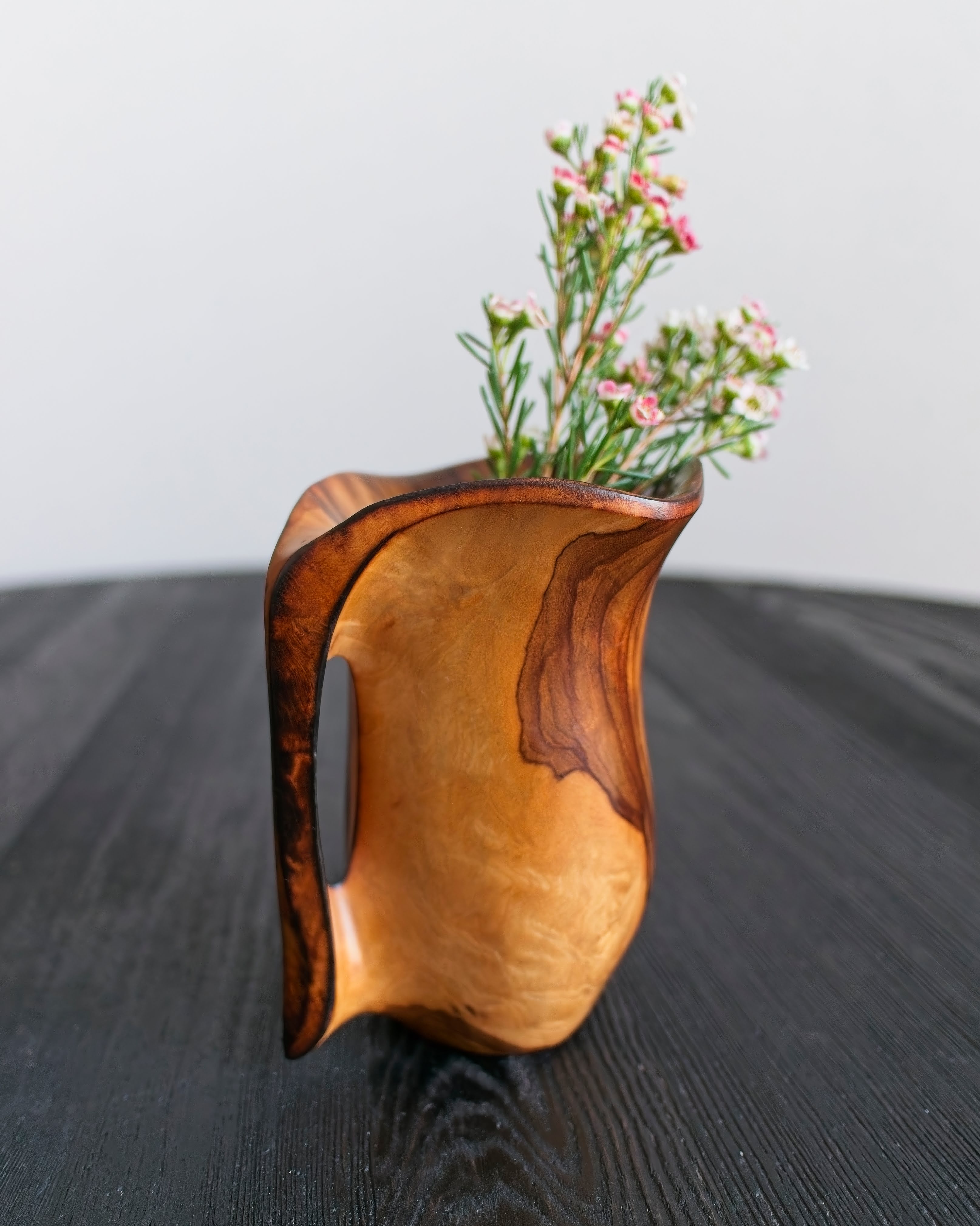 A Spanish vintage folk art olivewood vase with floral arrangement on a dark surface in front of a light grey background.