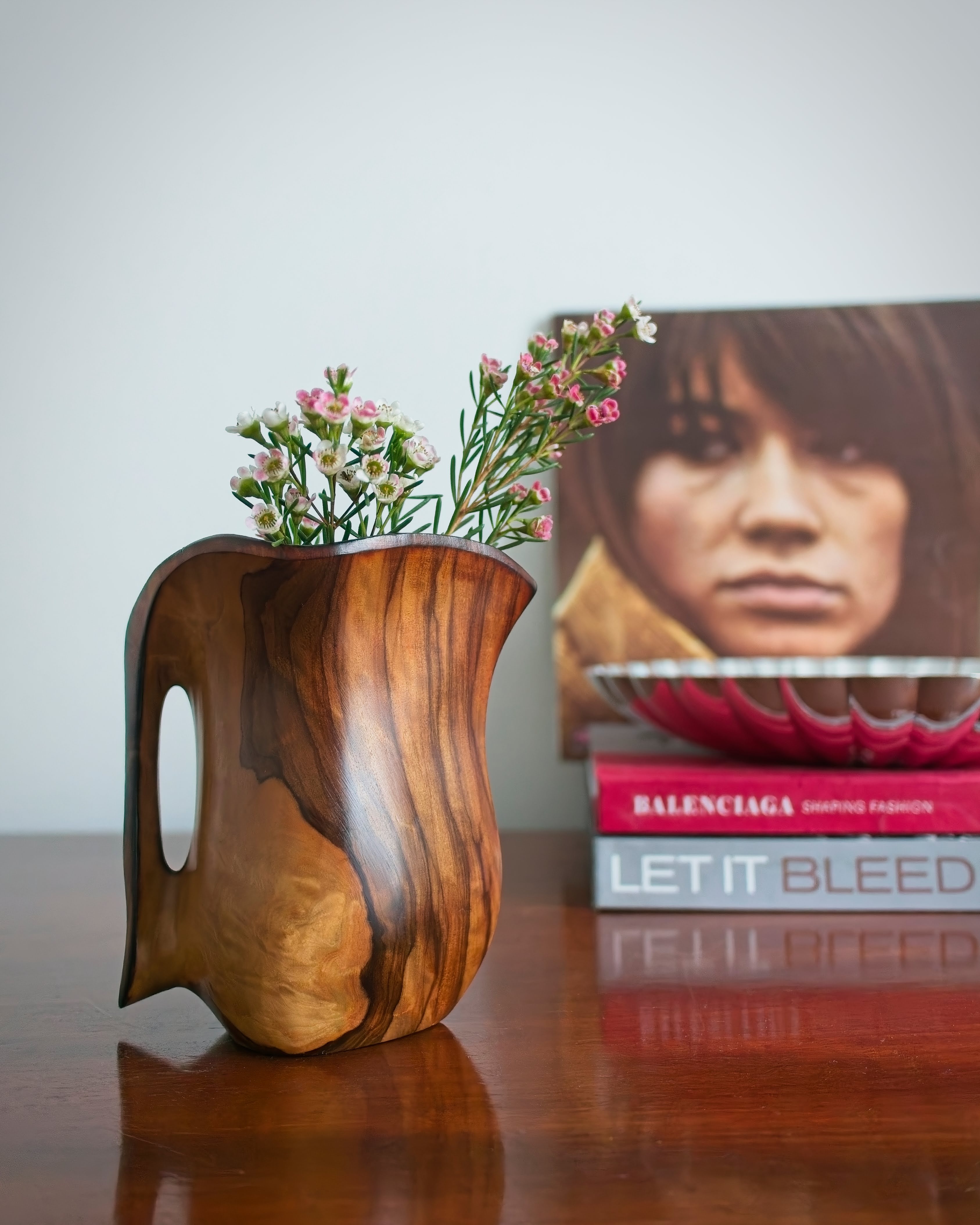A vintage wooden carved pitcher with intricate grain pattern containing small white flowers in front of a stack of books and a vintage vinyl record featuring the portrait of a young woman on the cover.