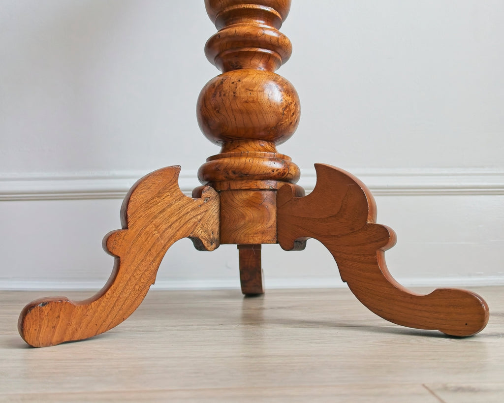 A close up of the pedestal base of a vintage maple burl wood accent table with a decorative design on a light wooden floor.