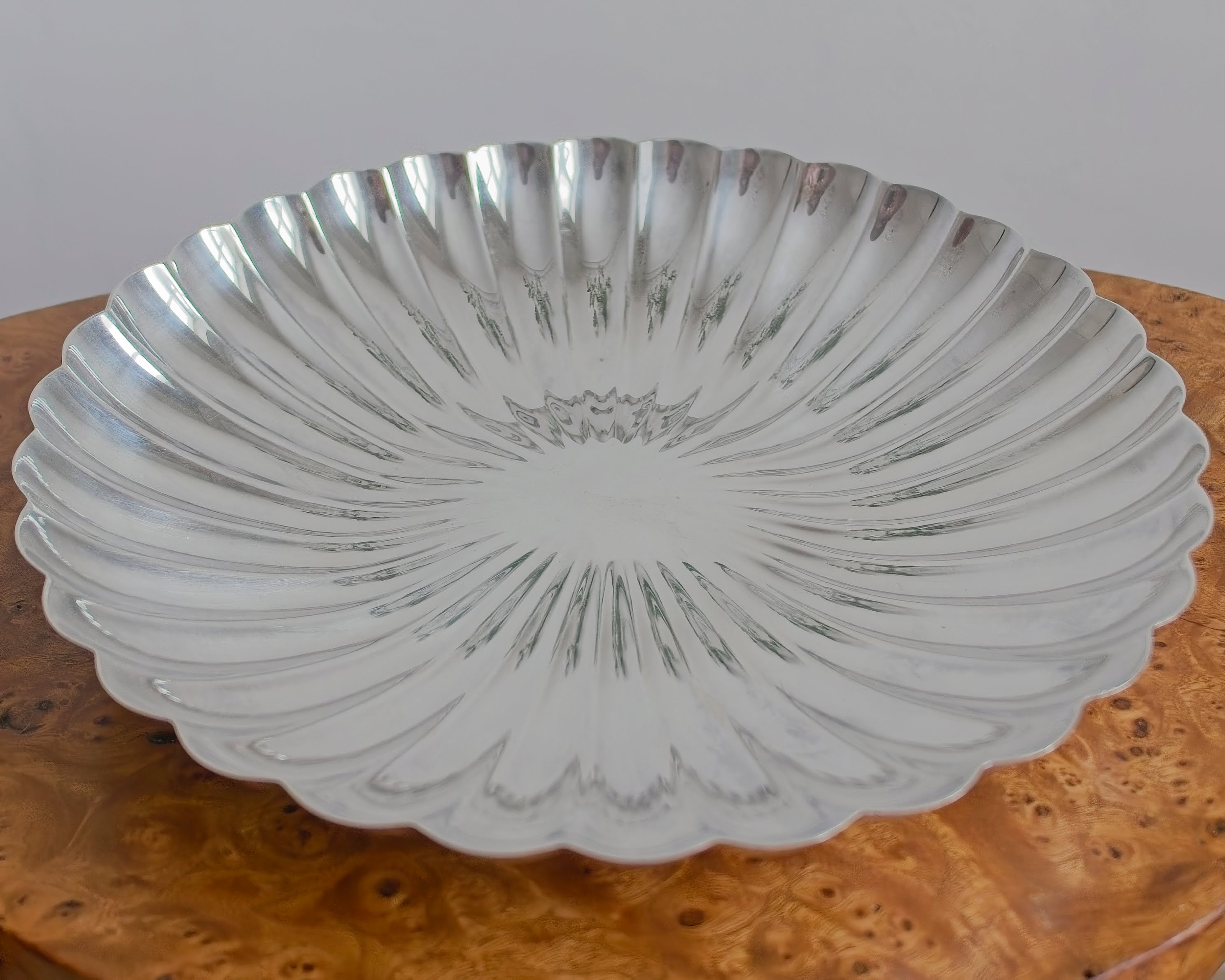 A close up of a vintage scalloped silver decorative bowl on a burr wood table.